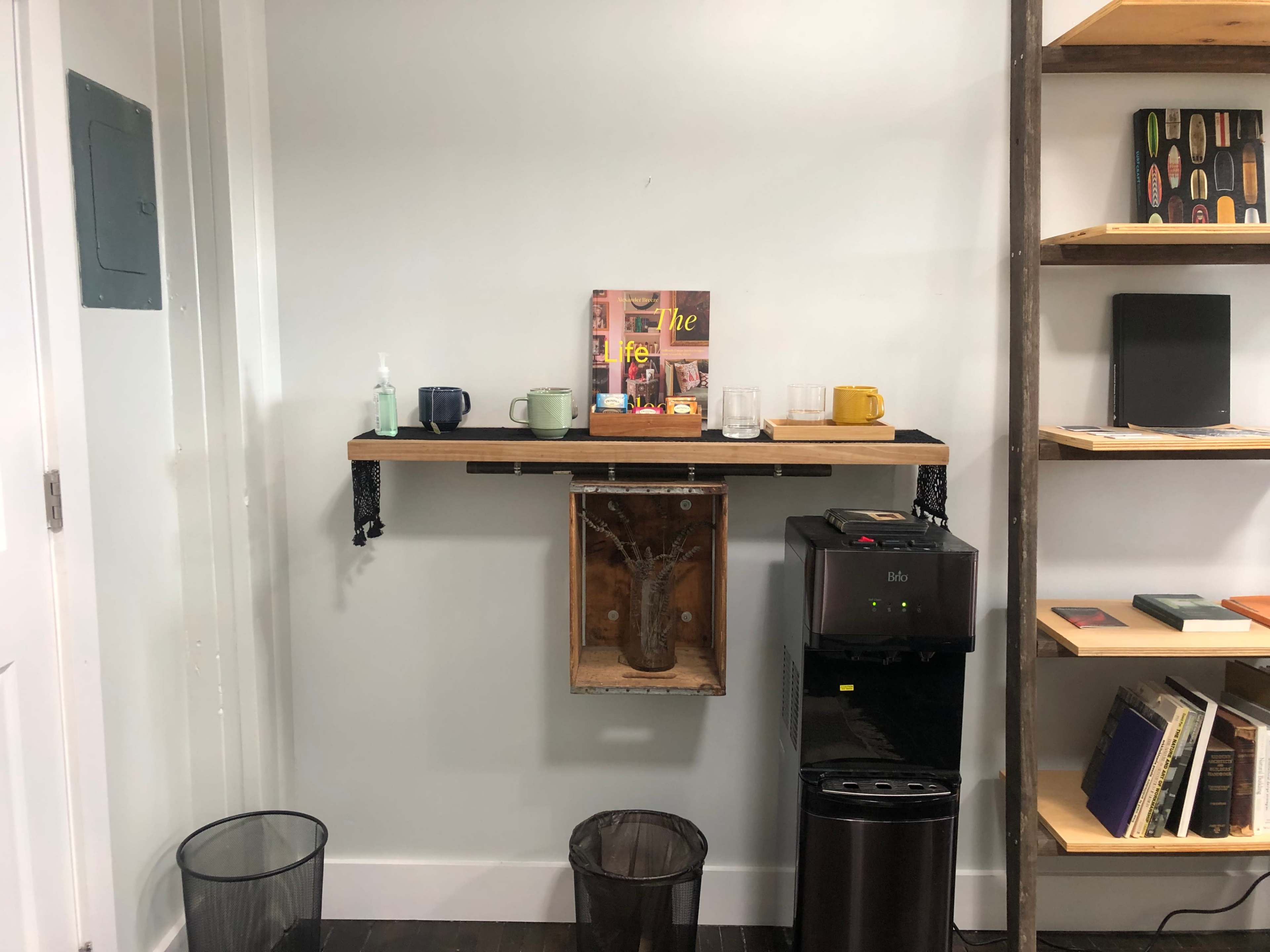 A wall-mounted shelf holds several mugs and glasses above a small refrigerator and two waste bins.