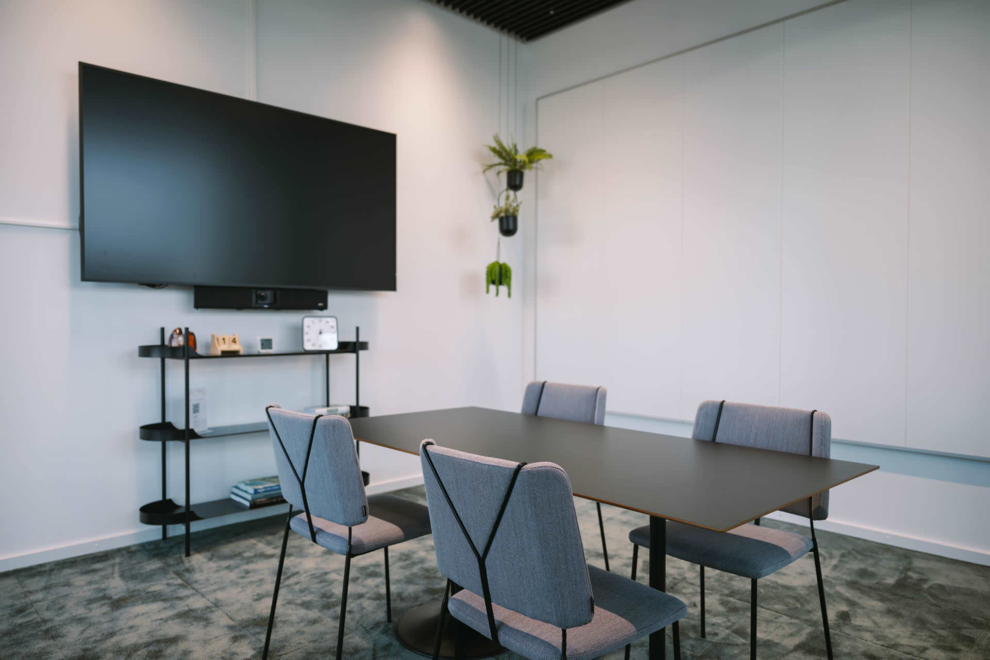 A modern conference room features a black table surrounded by four gray chairs, with a large wall-mounted television and a shelf displaying decorative items.