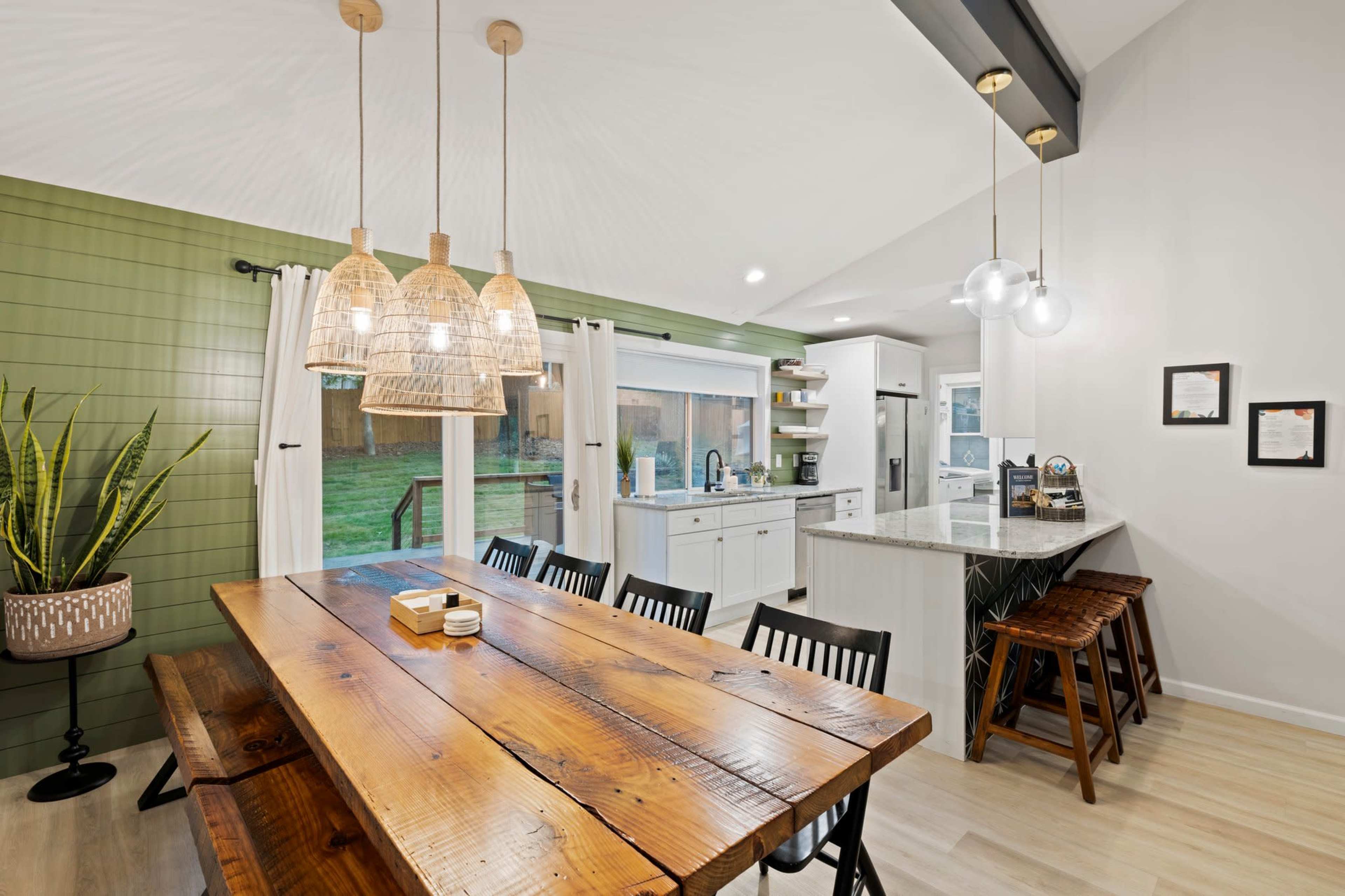 A bright kitchen features a large wooden dining table, modern pendant lights, and a minimalist design with green and white accents.