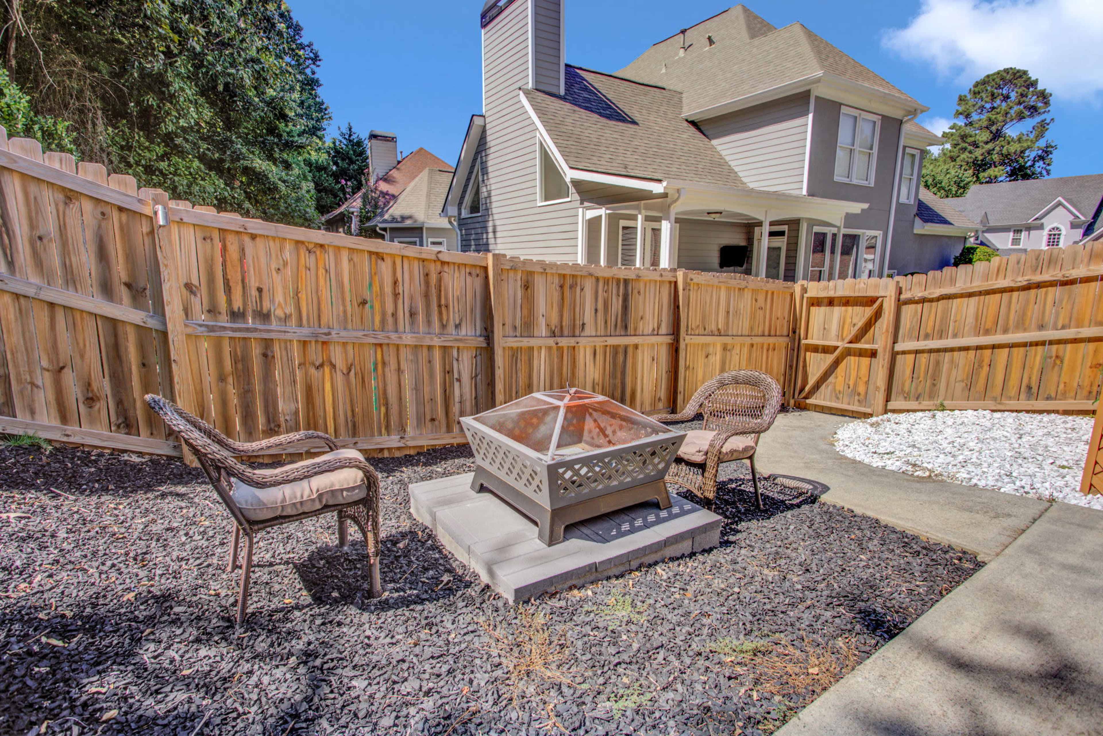 A small outdoor seating area surrounded by a wooden fence, featuring two chairs and a fire pit on a stone patio.