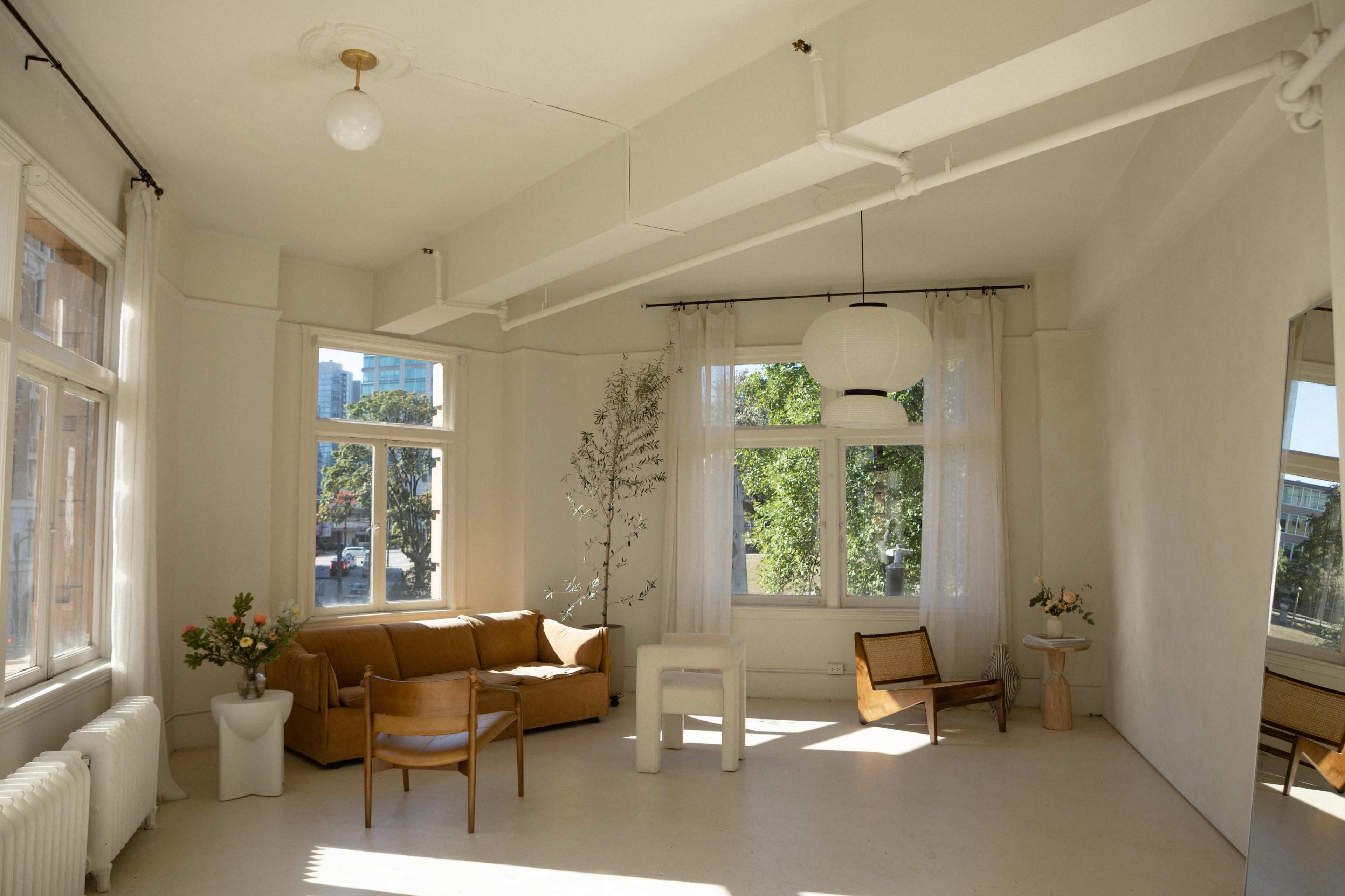 A bright, minimalist living space with large windows, a beige couch, a wooden chair, a small table with a vase, and light-colored walls.