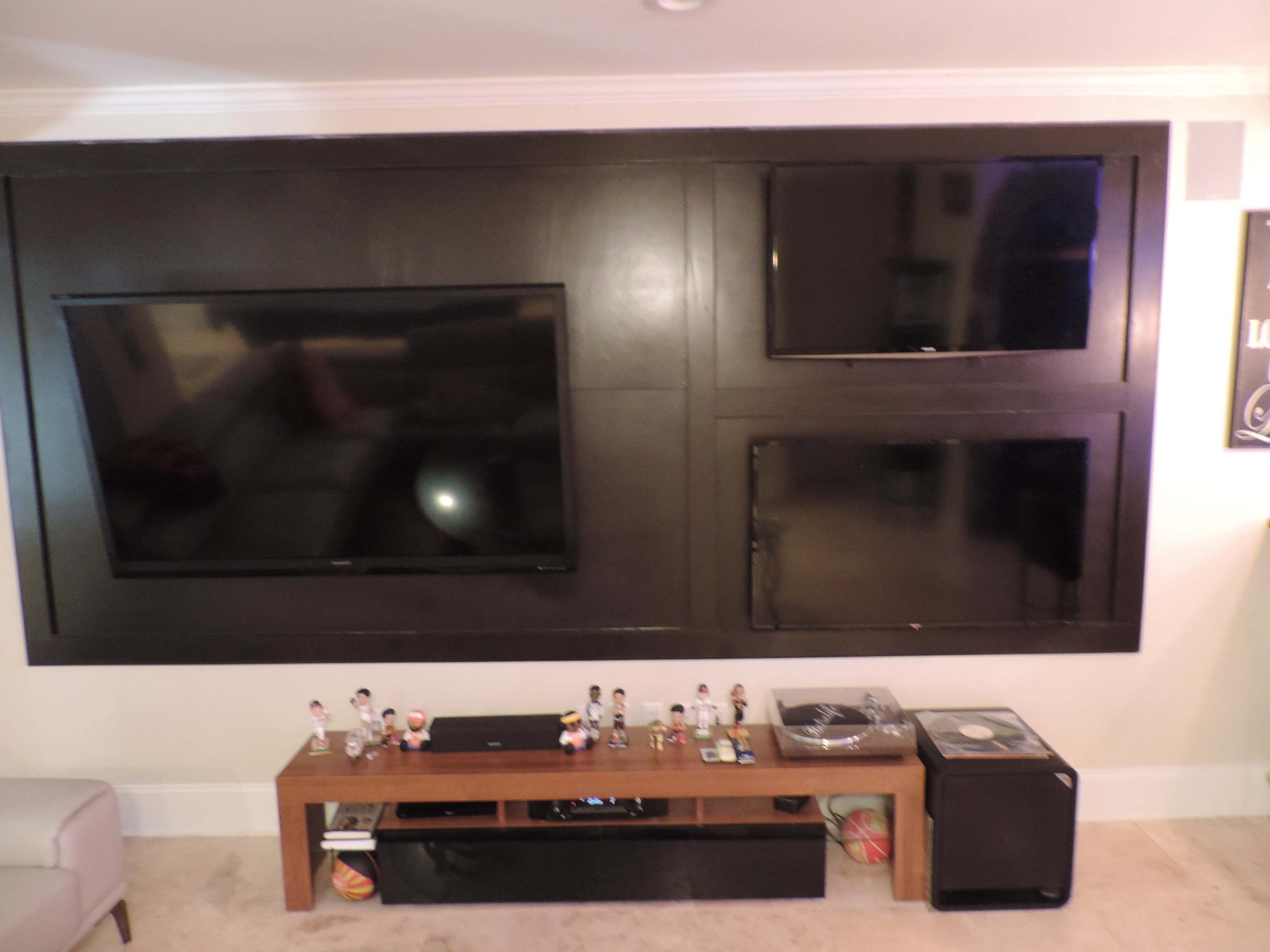 The image shows a living room with two mounted flat-screen TVs on a dark wood panel wall above a low wooden media console, which holds various items including a record player and small figurines.