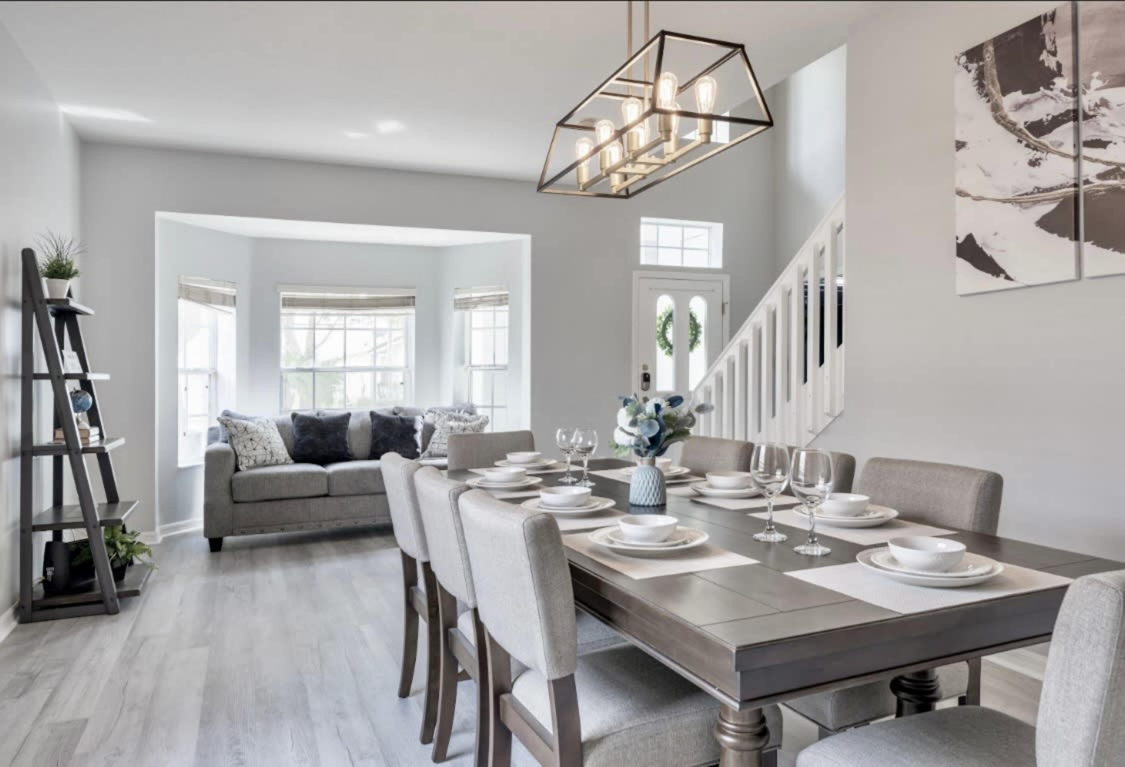 A modern dining room features a large table set for dinner, surrounded by beige chairs, with a cozy seating area visible in the background.