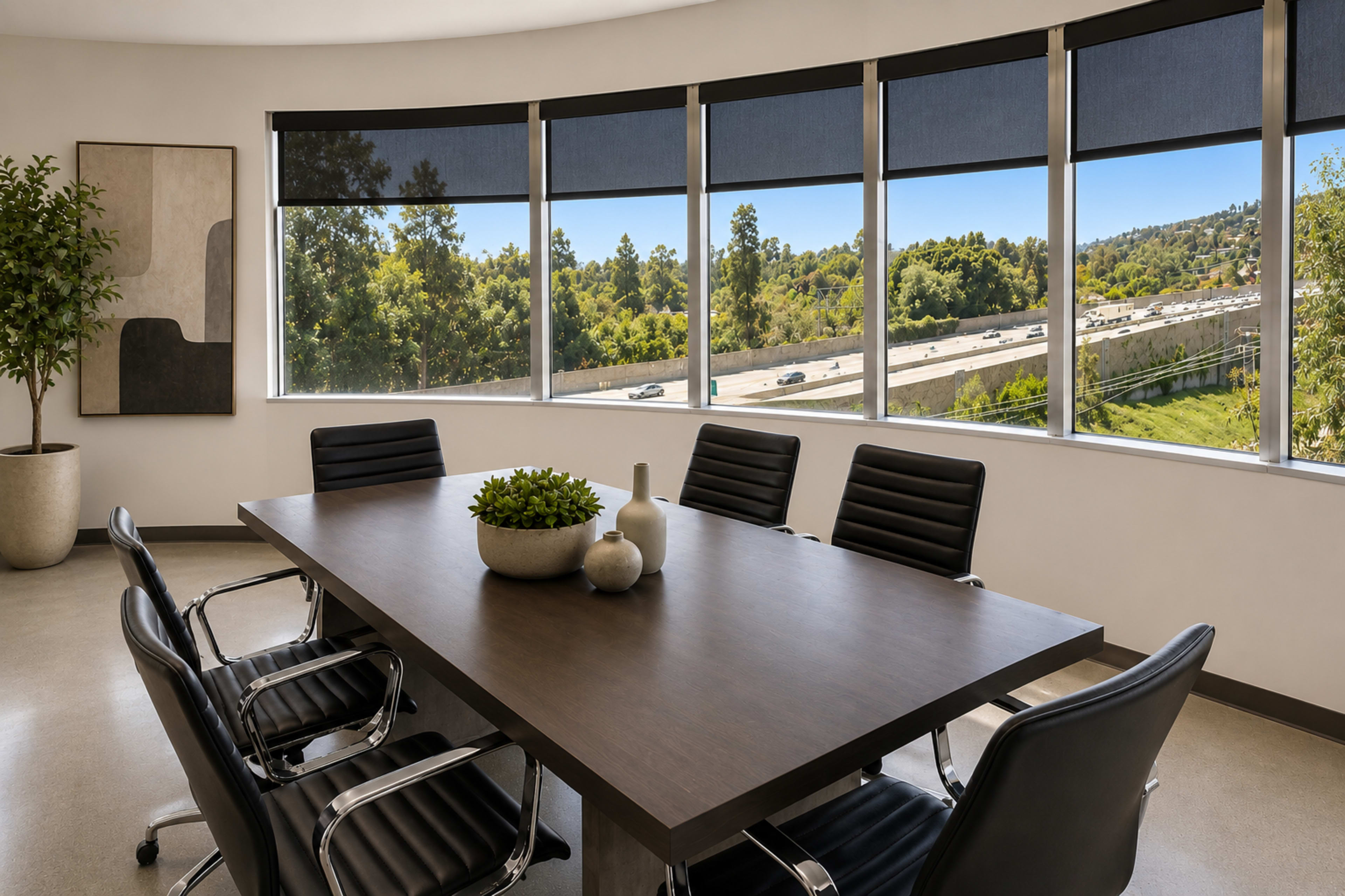 A conference room features a large wooden table surrounded by black chairs, with large windows providing a view of a tree-lined highway.