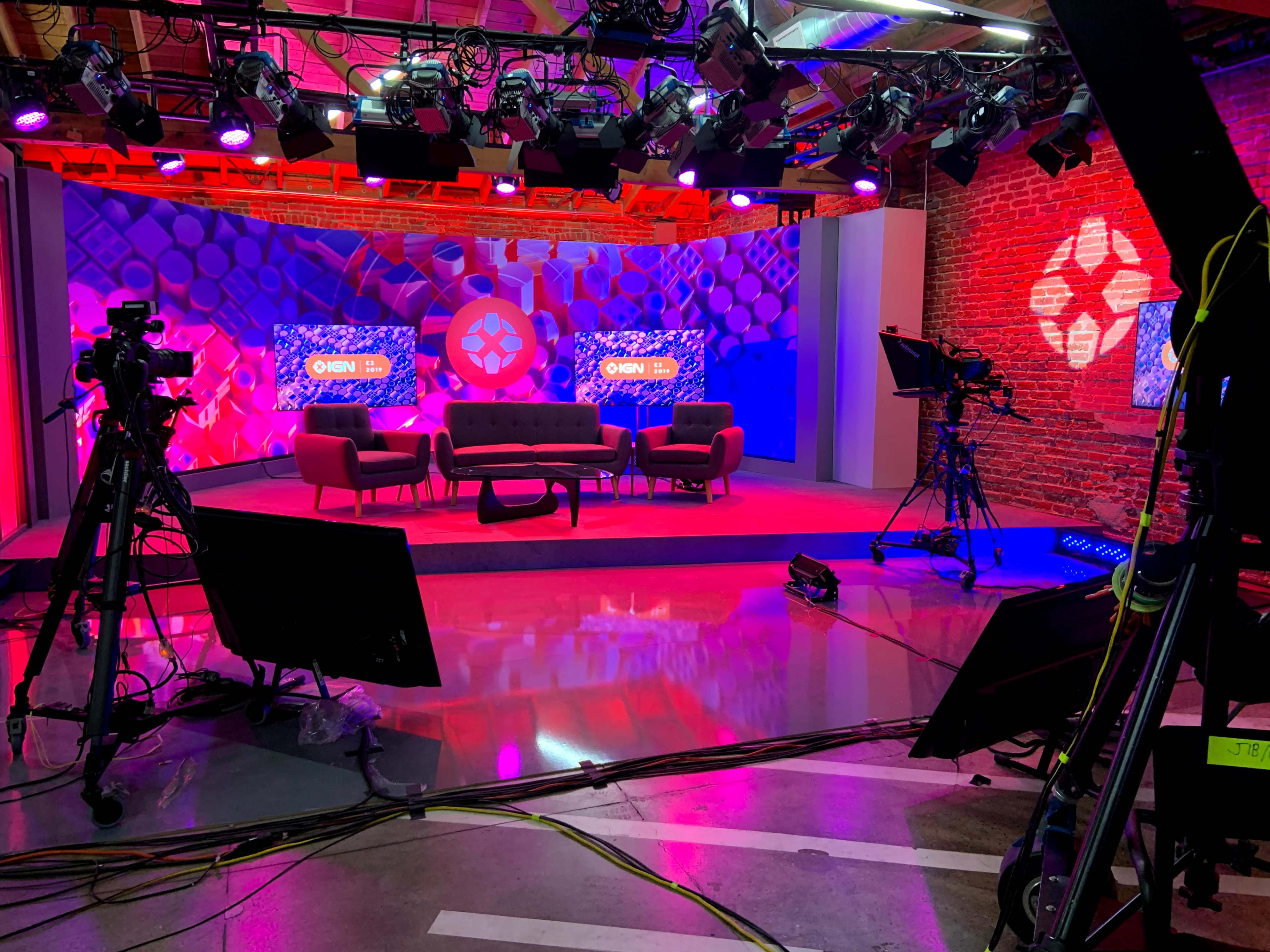 A TV studio set features a lounge area with a sofa and colorful lighting, surrounded by cameras and production equipment.
