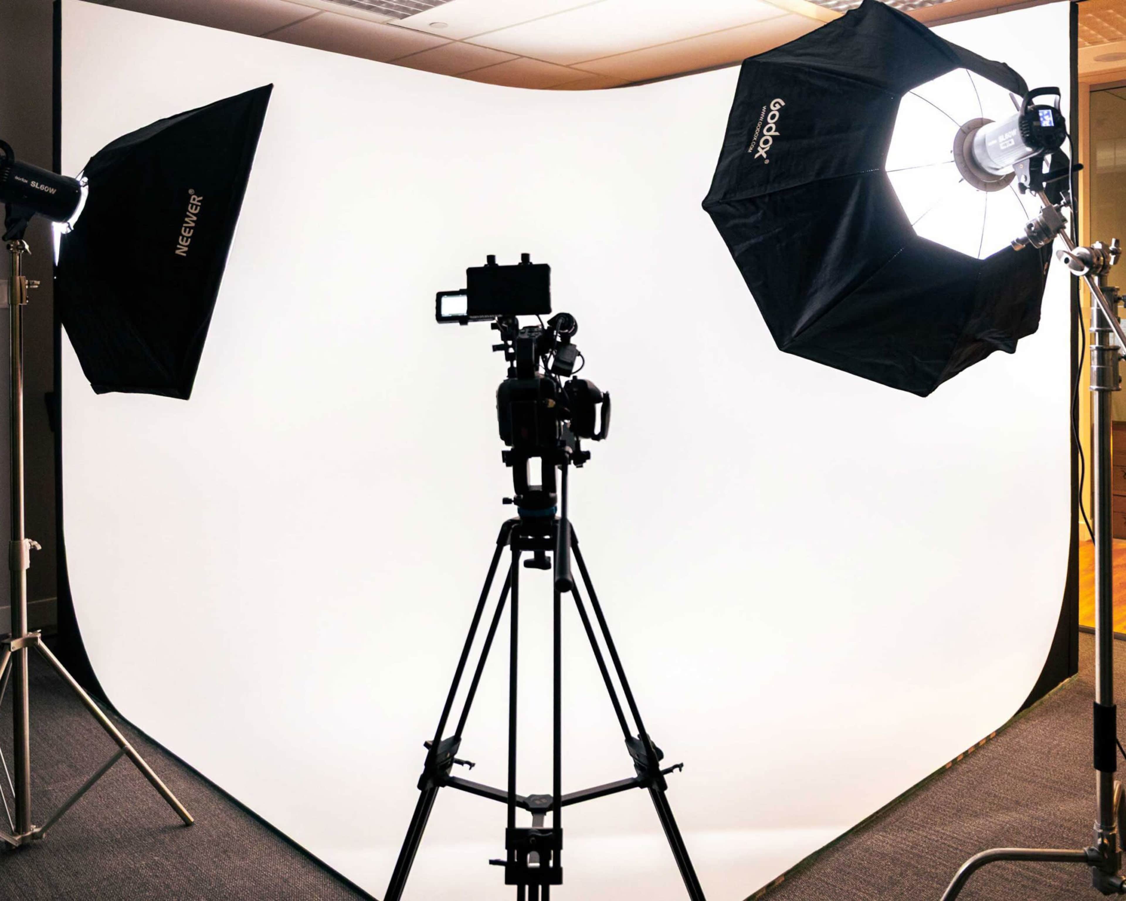 A photography studio setup features a camera on a tripod with two softbox lights illuminating a white backdrop.