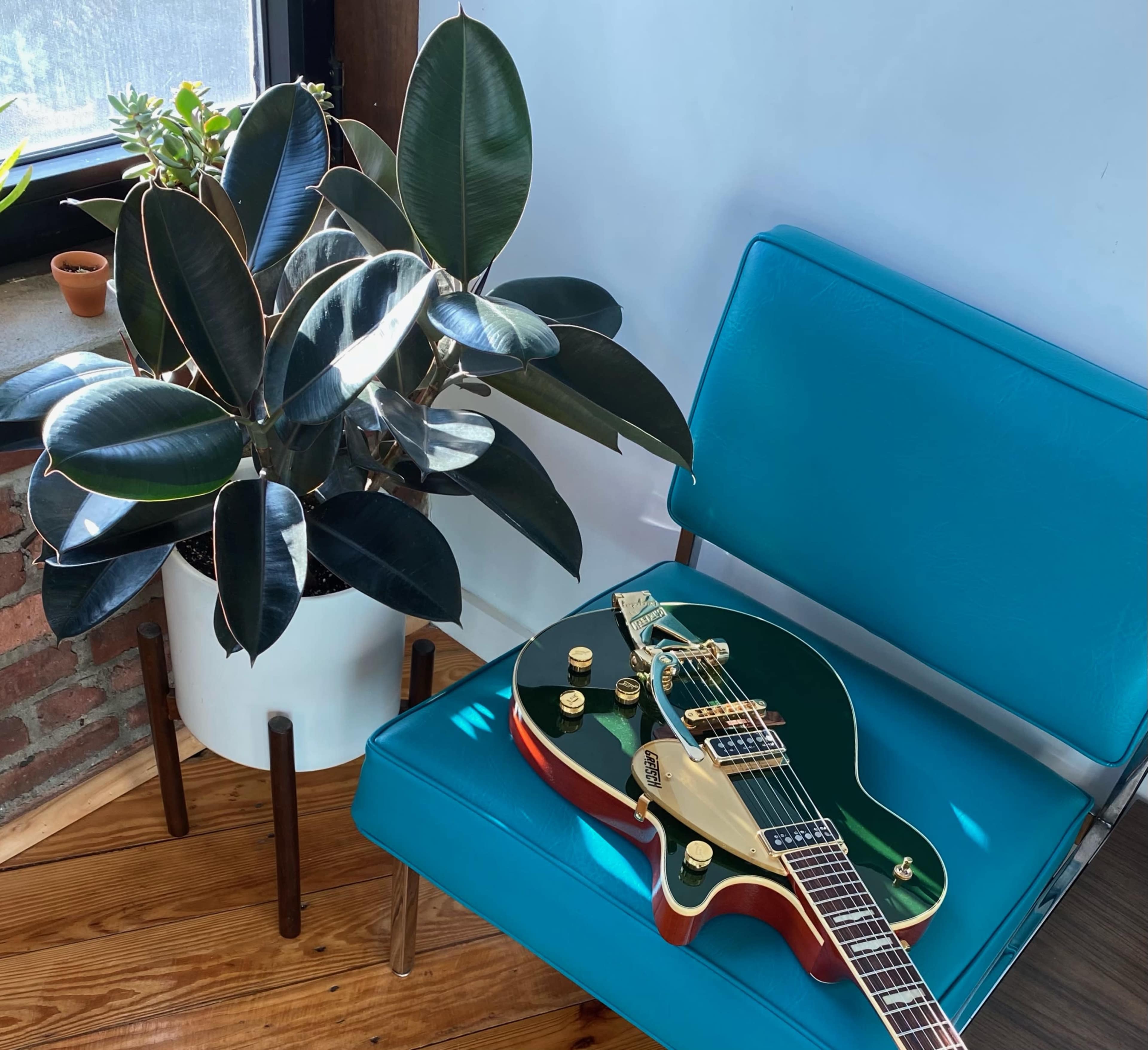 A green electric guitar rests on a bright blue chair beside a large potted plant in a well-lit room.