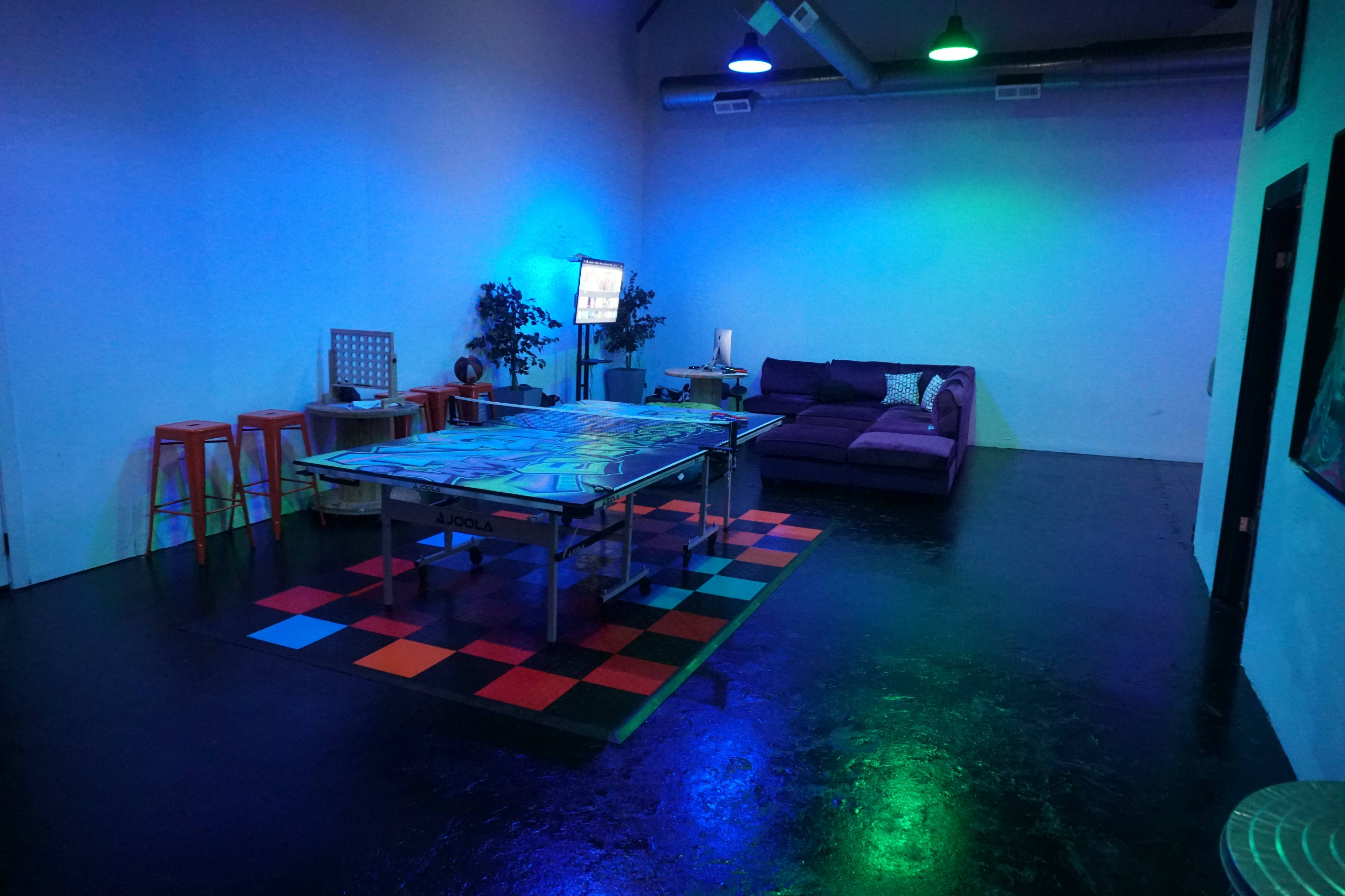 The room features a ping pong table on a colorful checkered mat, with a purple couch and bar stools against the walls, illuminated by blue and green lighting.