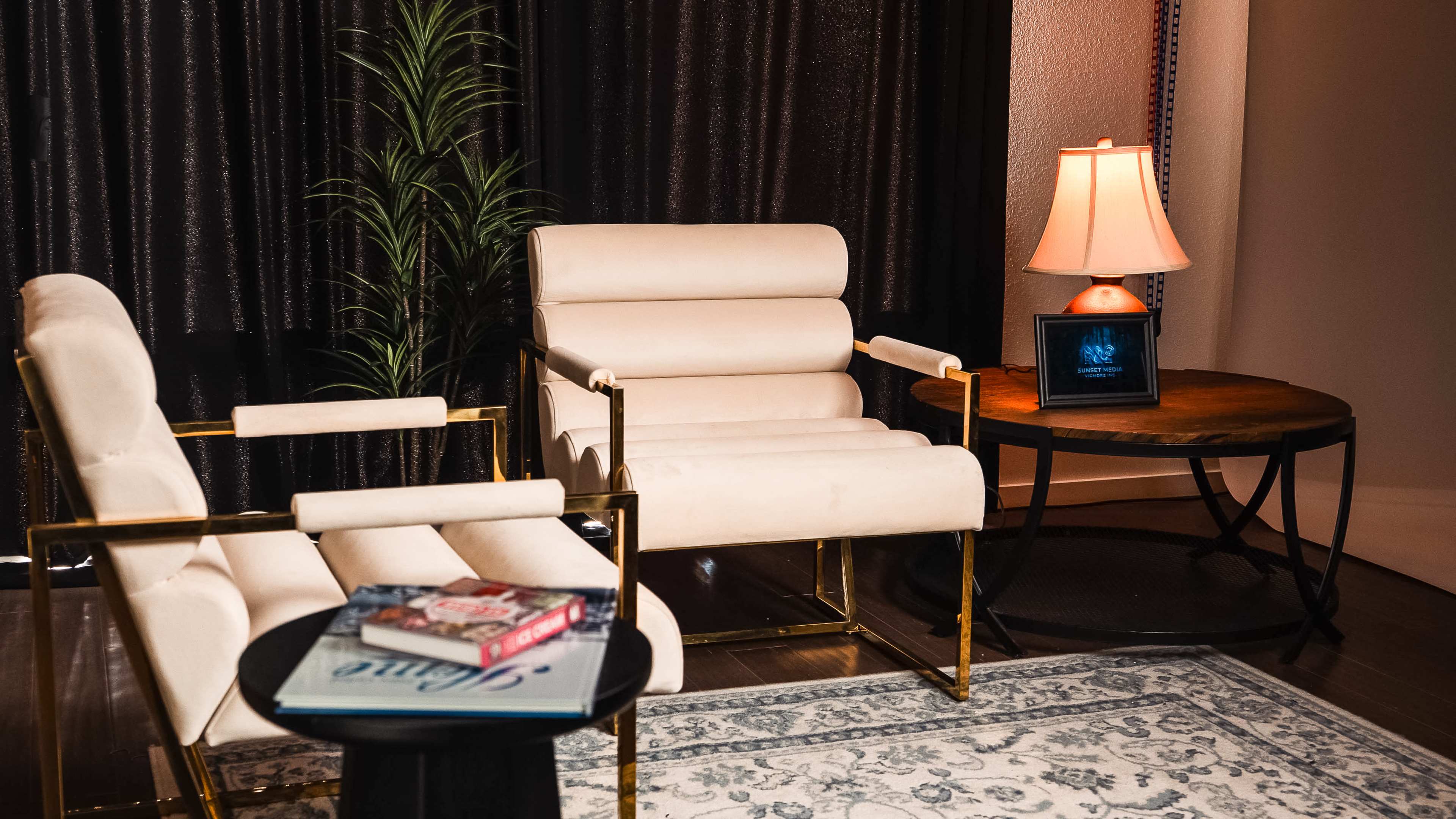 The image shows a cozy seating area with two cream-colored chairs, a round black table with books on it, a small lamp, and a potted plant against a dark backdrop.