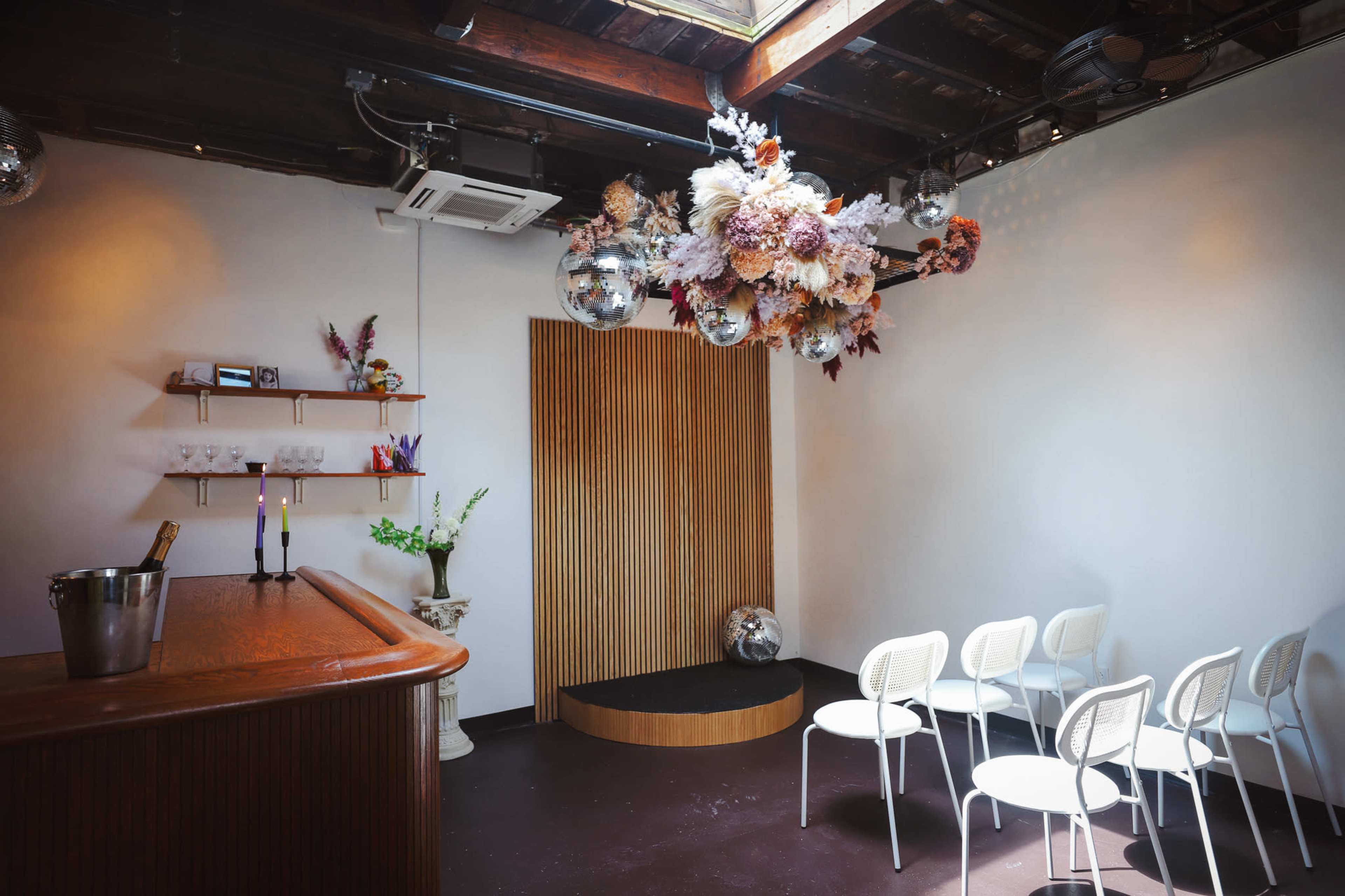 A stylish interior space featuring a wooden bar, a flower and disco ball installation overhead, and a small stage area with white chairs arranged in front of it.