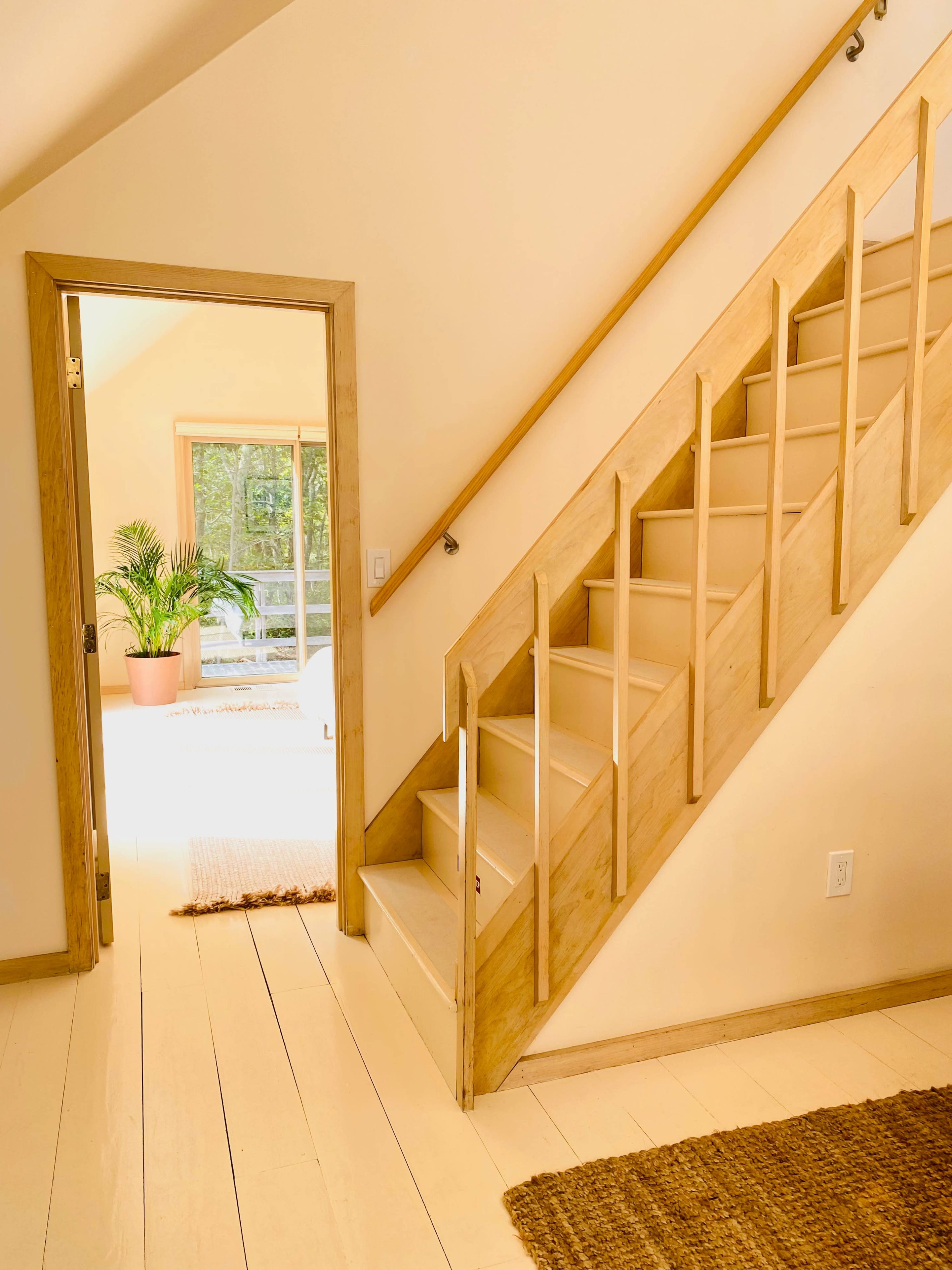 The image shows a staircase leading to an upper level, with an open door revealing a bright room and a potted plant.