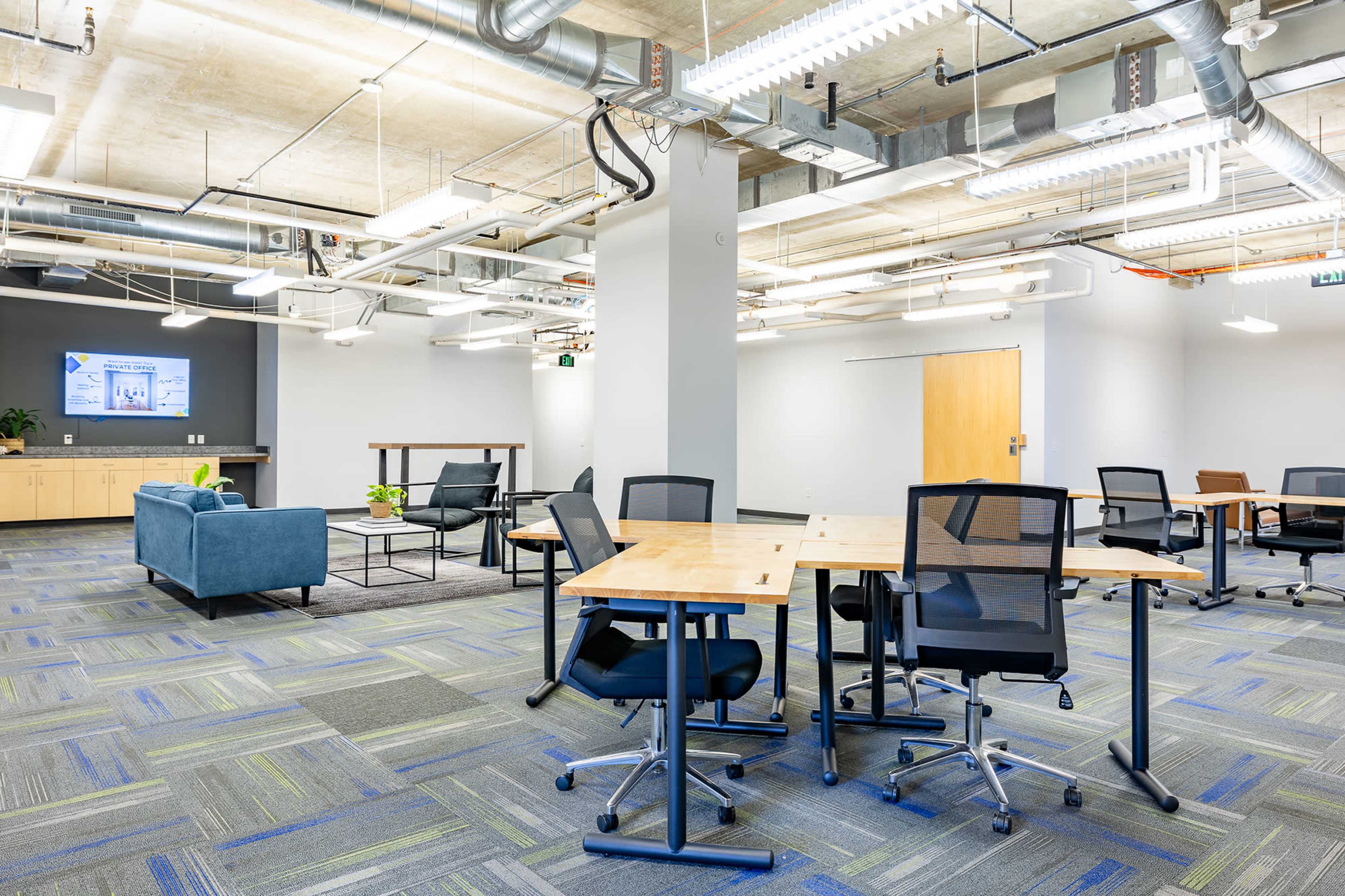 The image shows a modern office space with tables and chairs arranged for collaboration, featuring gray walls, a television screen, and a seating area with a blue couch.