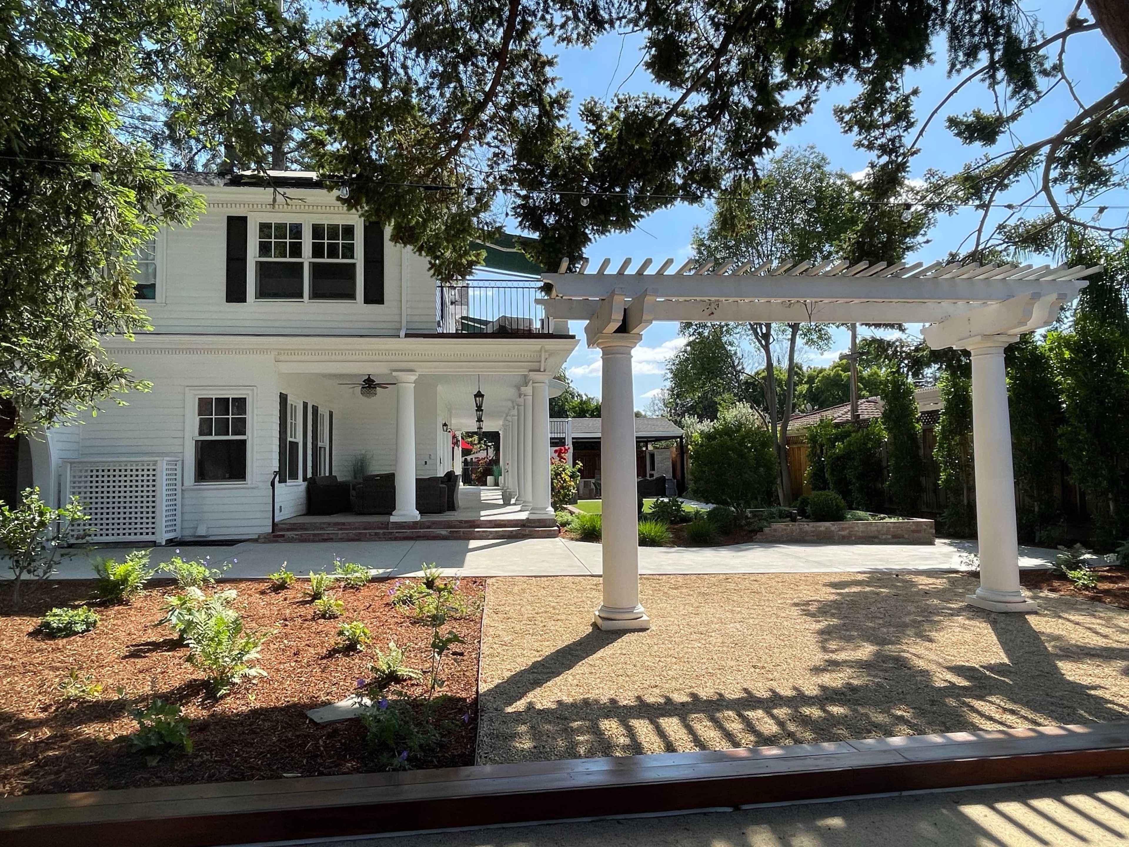 A two-story white house with a pergola and a landscaped yard features a gravel area surrounded by plants.