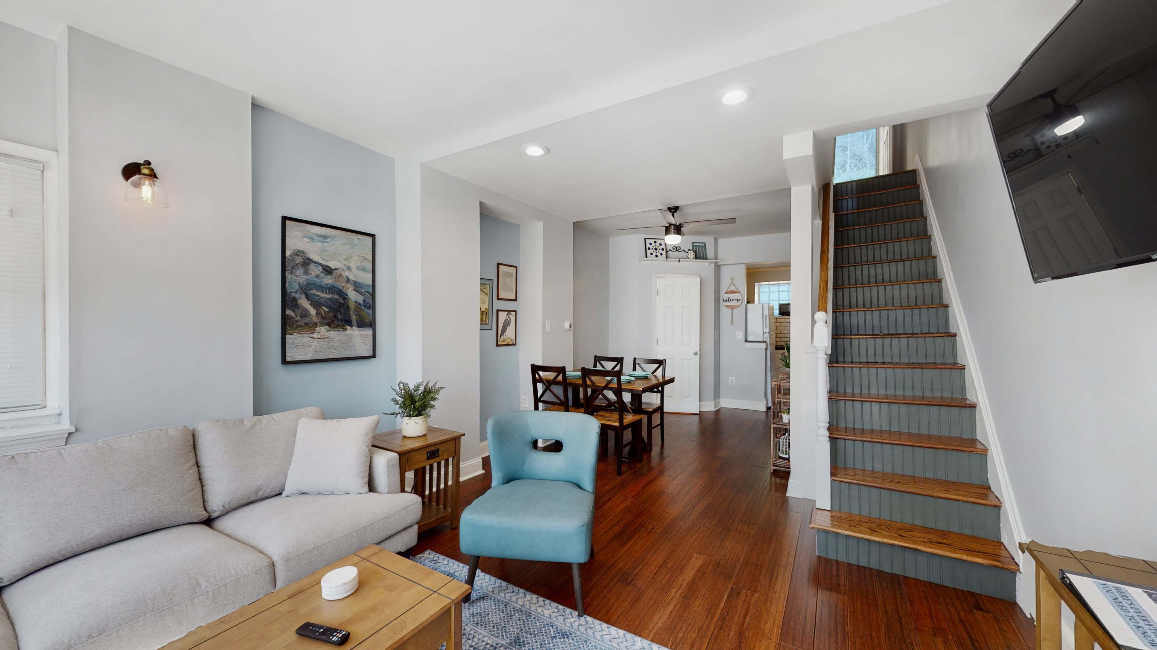 A modern living room with a couch, chair, and wooden table, leading to a dining area and a staircase.