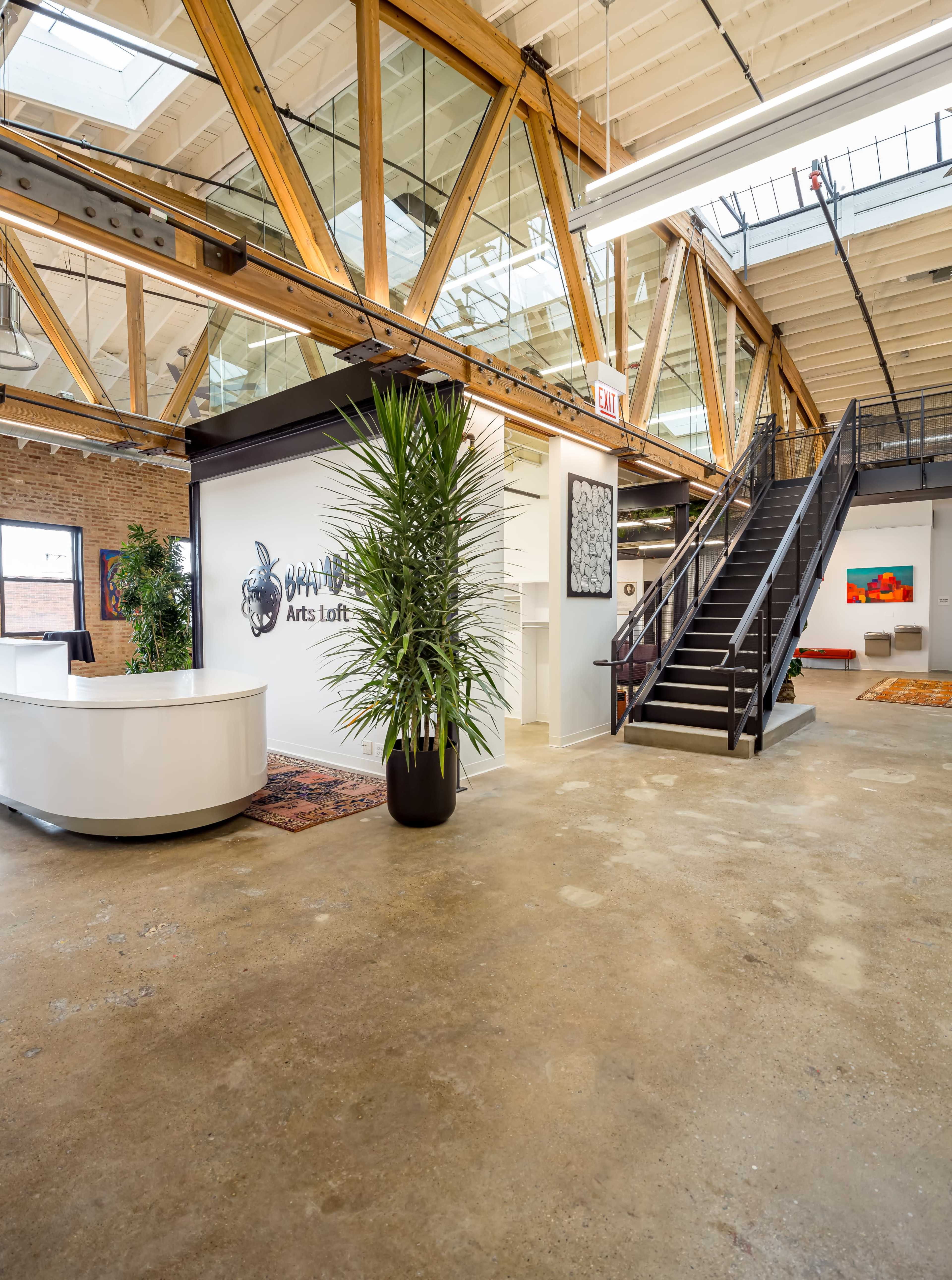 A spacious interior of an arts loft with wooden beams, a white reception desk, and a staircase leading to an upper level.