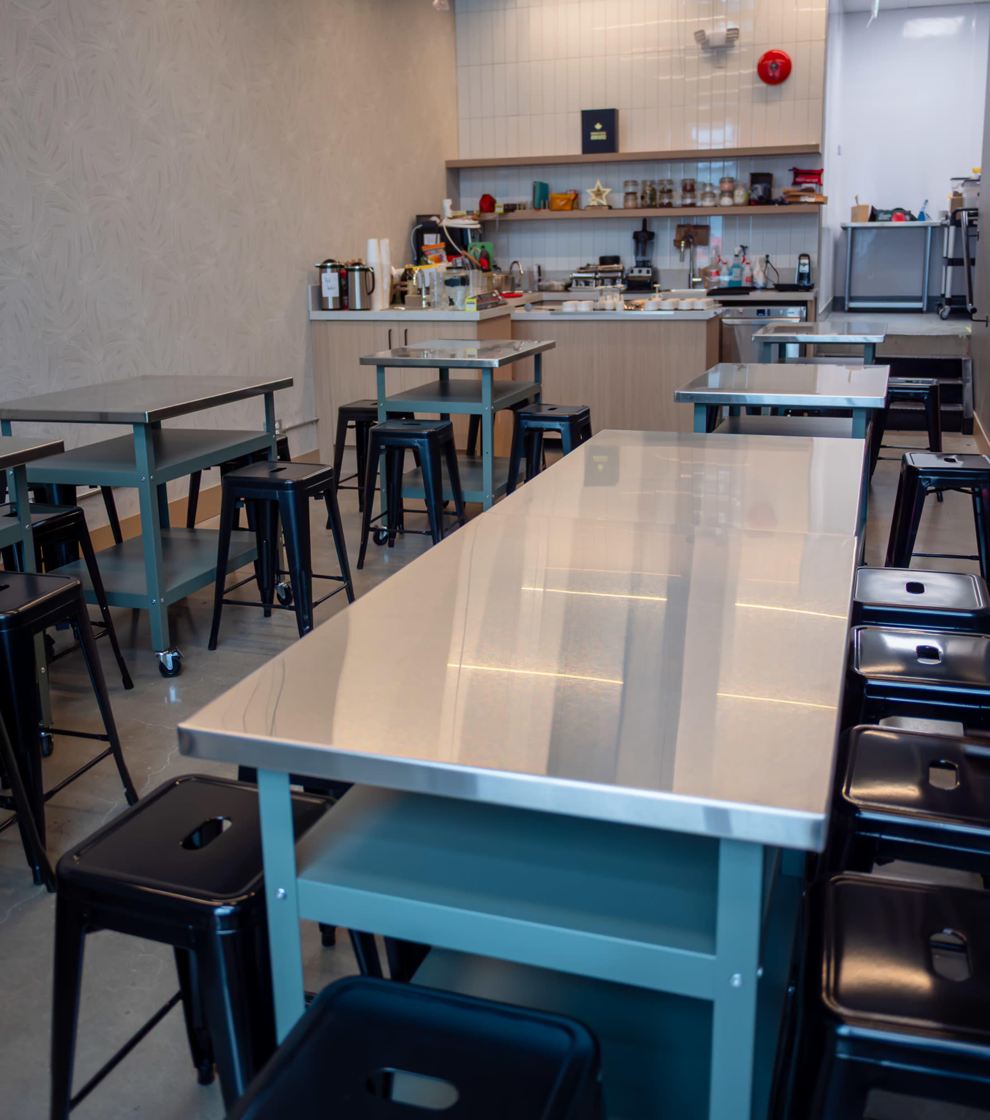 The image shows a modern, minimalist café or kitchen space with stainless steel tables and metal stools arranged neatly, featuring a counter and various kitchen items in the background.