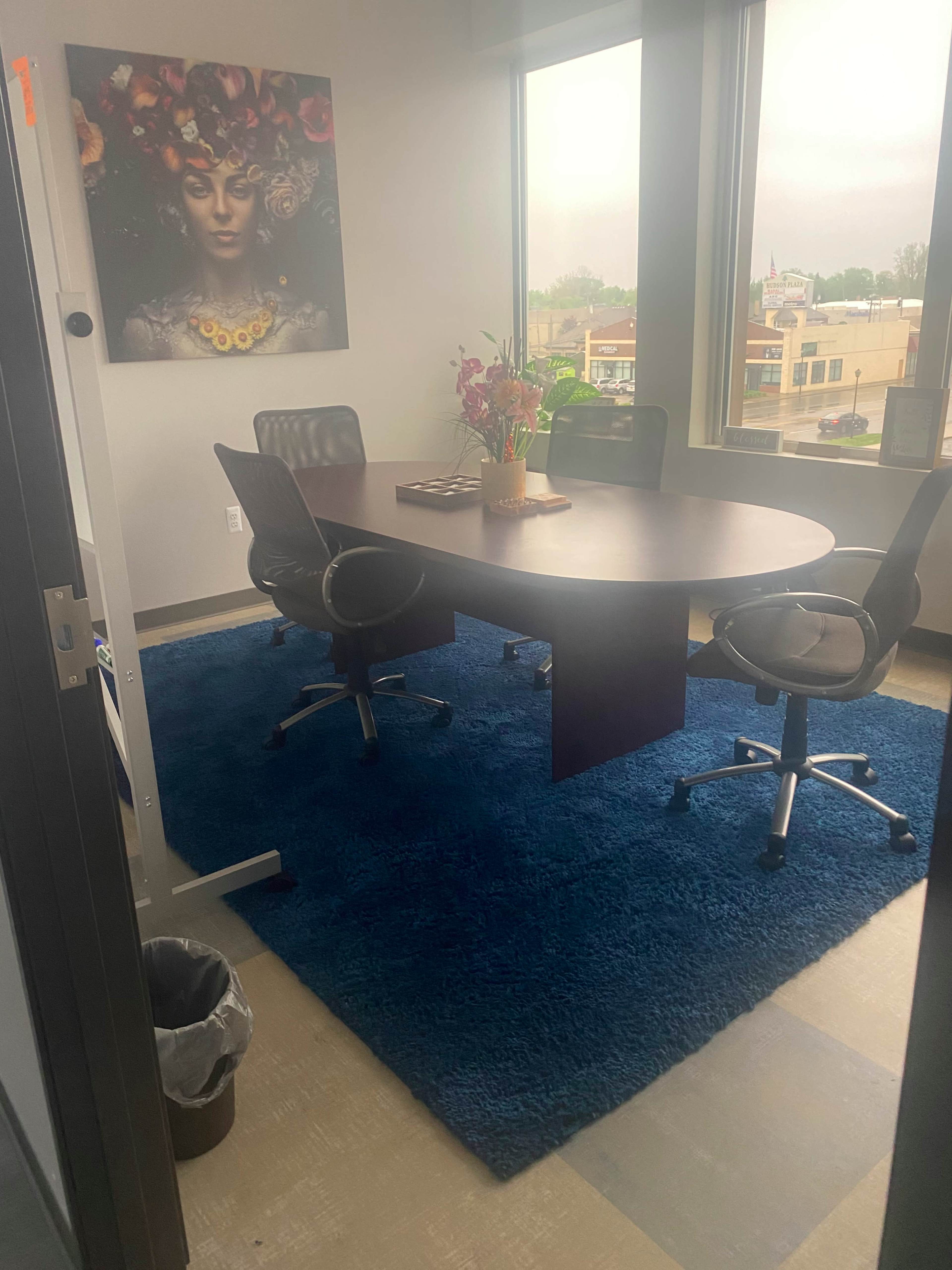 A conference room with a round table surrounded by four chairs, featuring a large window, a colorful wall art piece, and a rug on the floor.