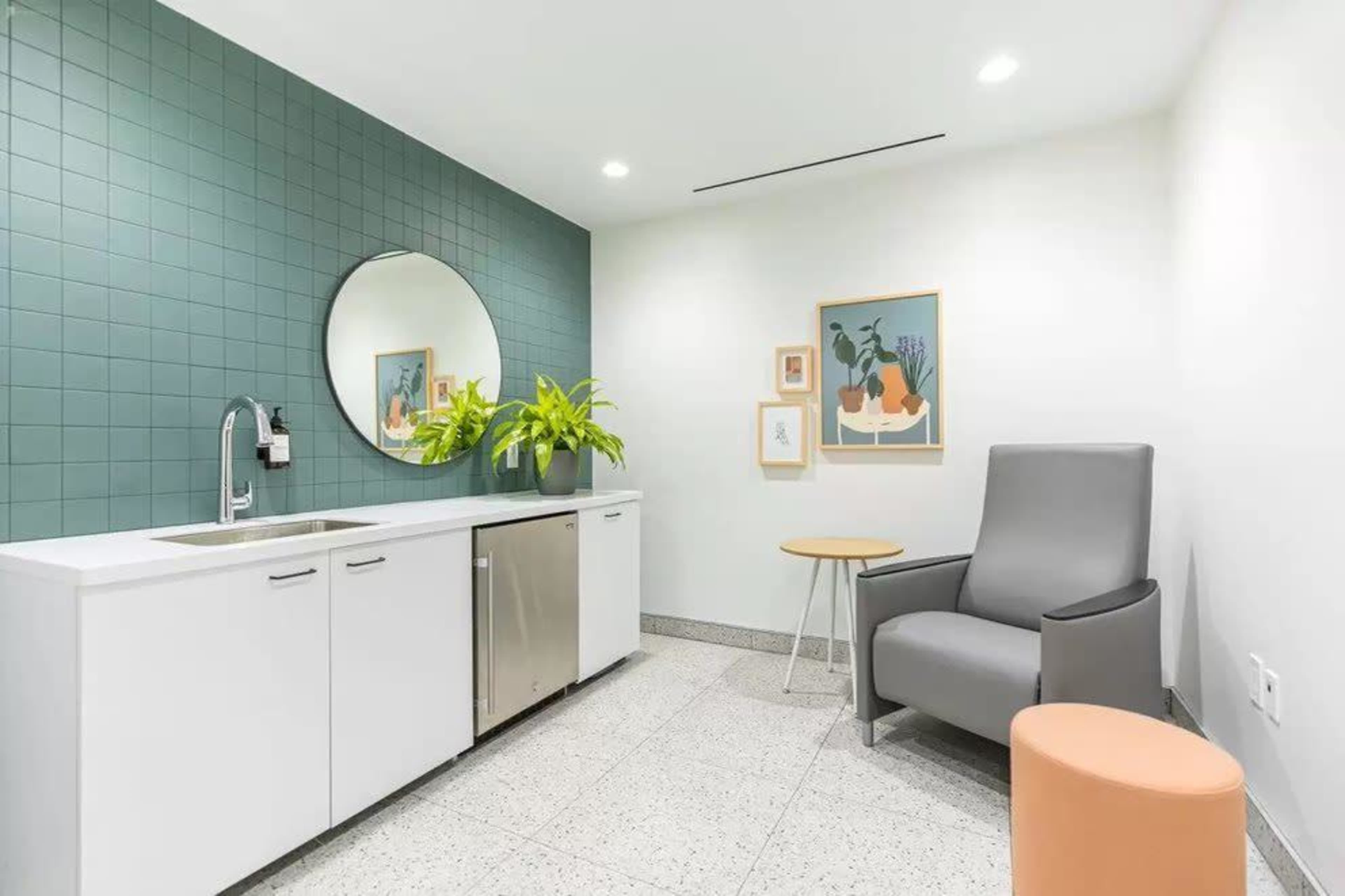 A modern break room with a green tiled wall, a sink, a small fridge, potted plants, a round mirror, framed artwork, a gray armchair, and a small round table.