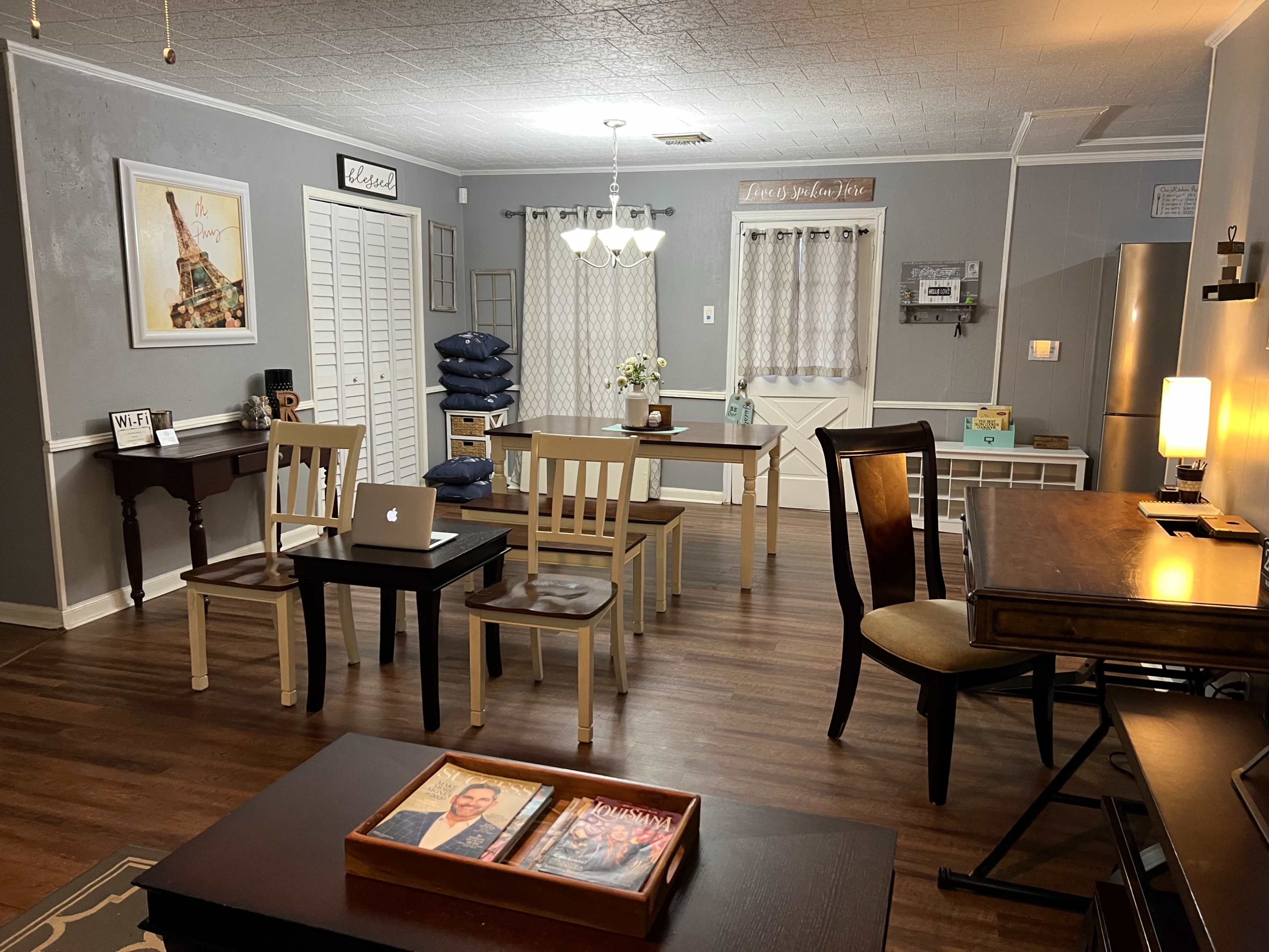 A spacious room with wooden flooring, featuring various tables and chairs arranged around, and a subtle decor with a few wall hangings and windows.