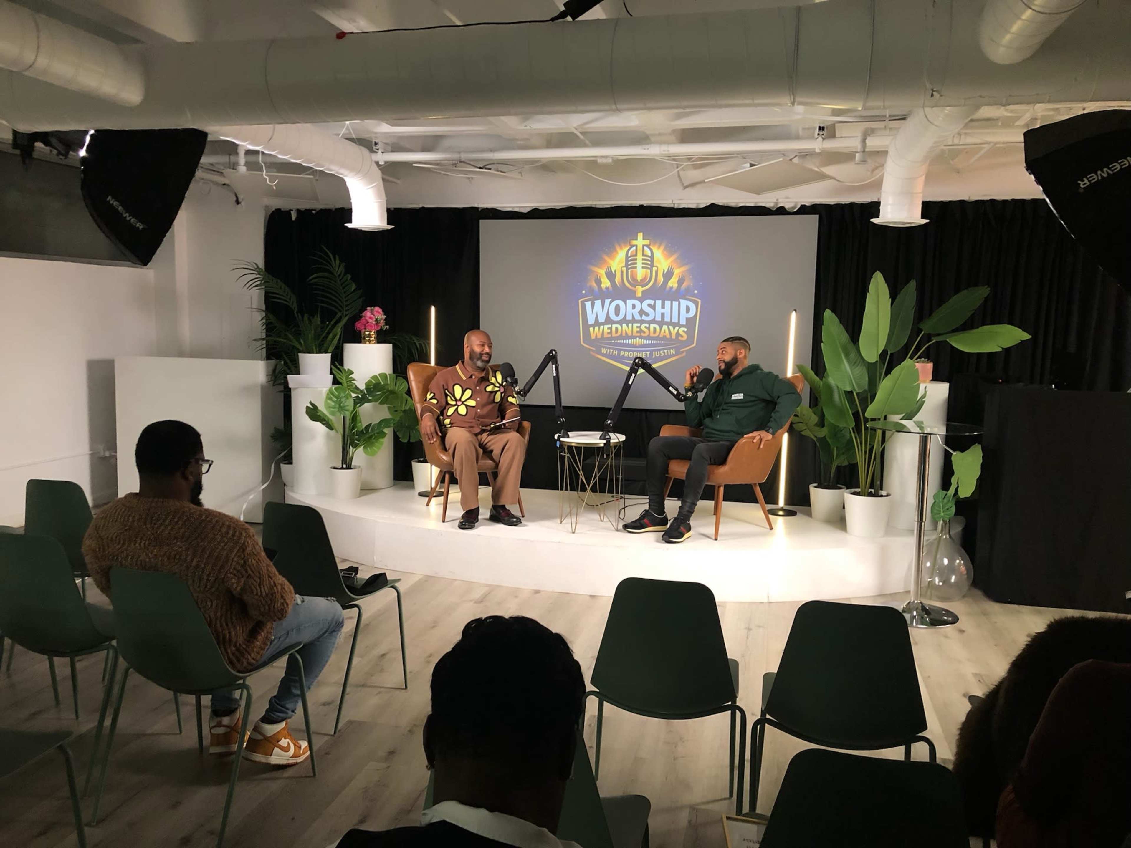 Two men are seated in stylish chairs on a stage with a decorative backdrop for an event labeled "Worship Wednesdays," while an audience sits in chairs facing them.