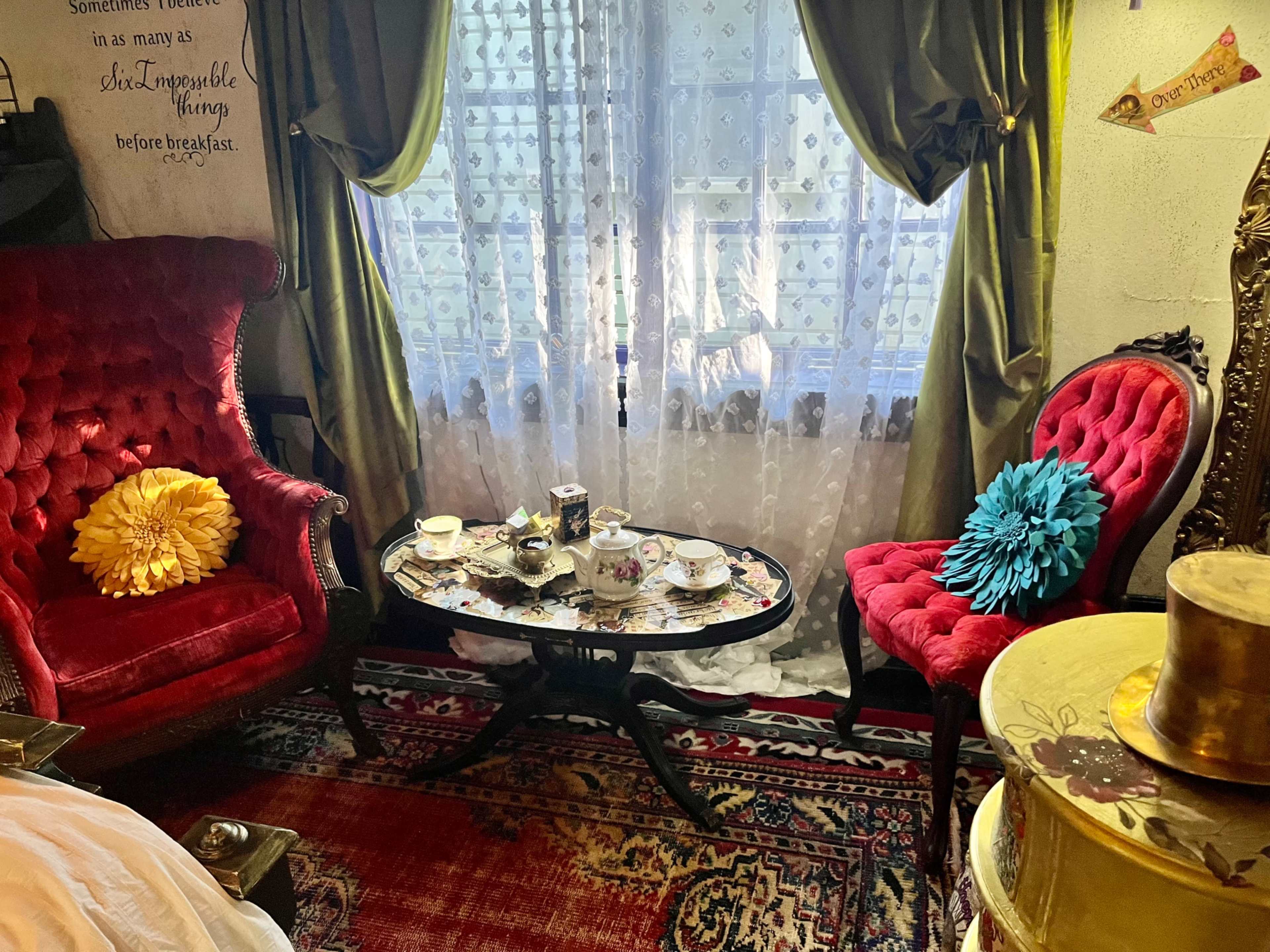 The scene depicts a quaint seating area with two red velvet chairs, a decorative coffee table covered in tea set items, and a window adorned with lace curtains.
