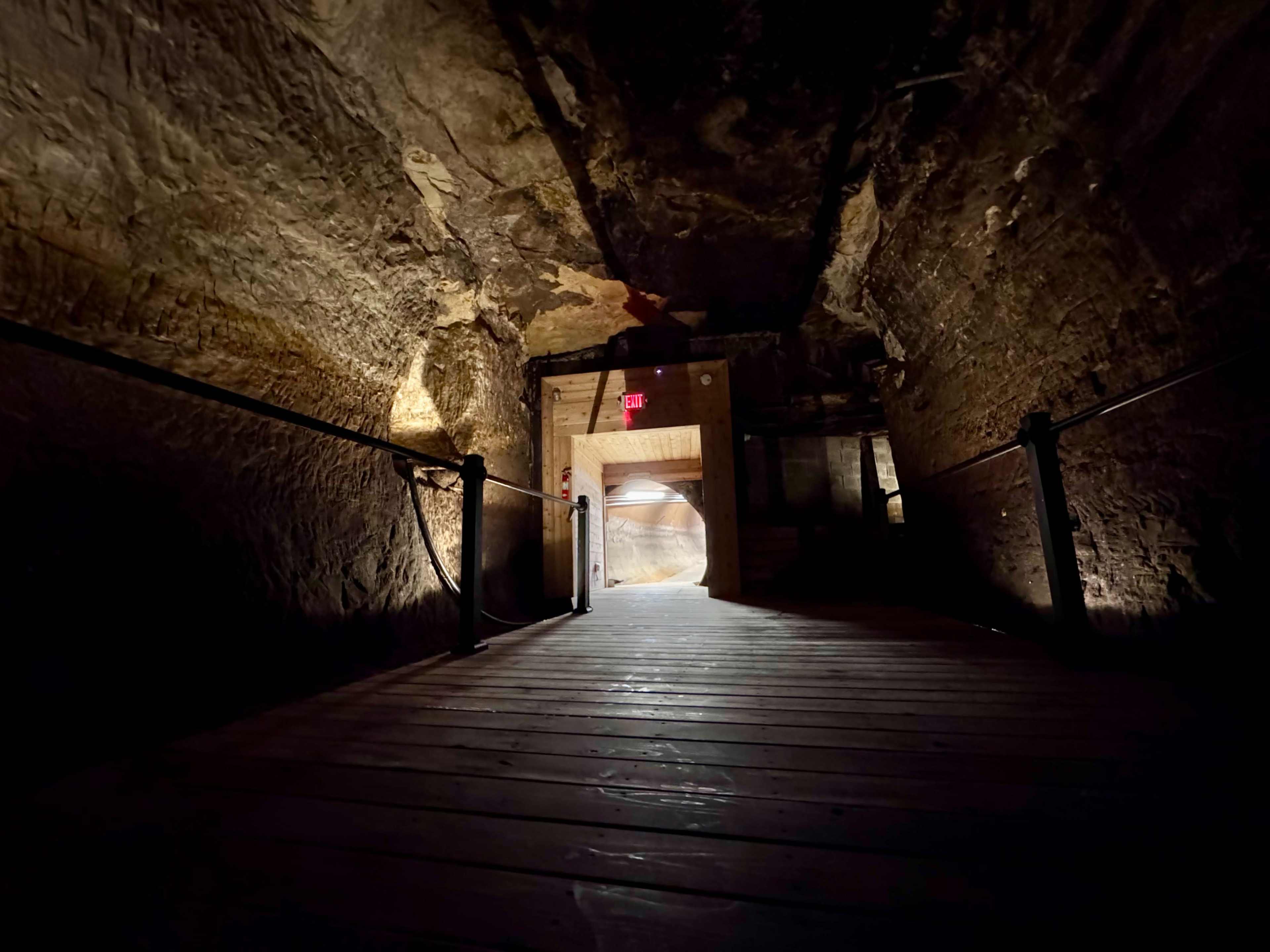 The image shows a dimly lit tunnel made of stone, leading to an arched opening illuminated by warm lights.
