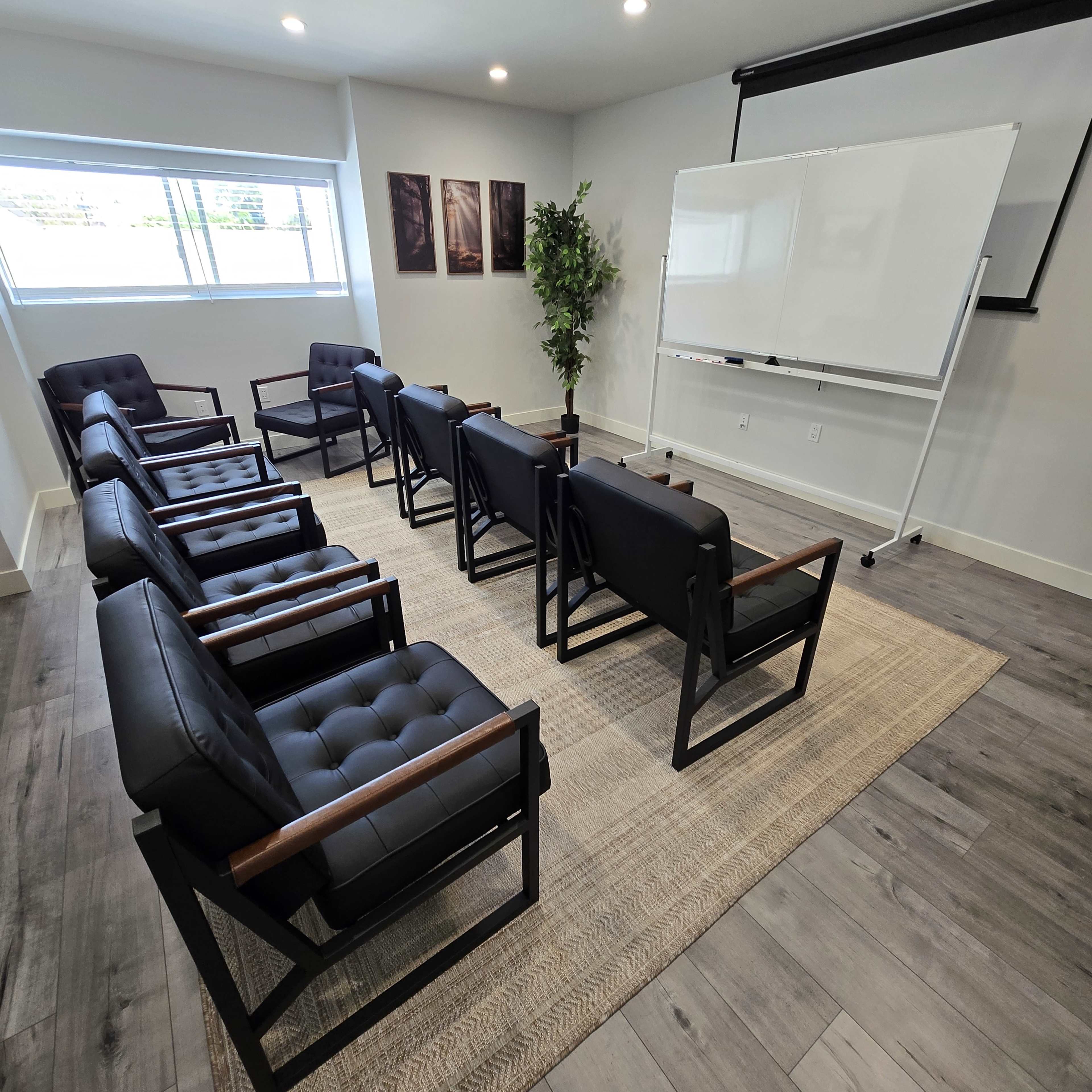 The image shows a modern meeting room with seven black chairs arranged in a circle around a large whiteboard and a potted plant.