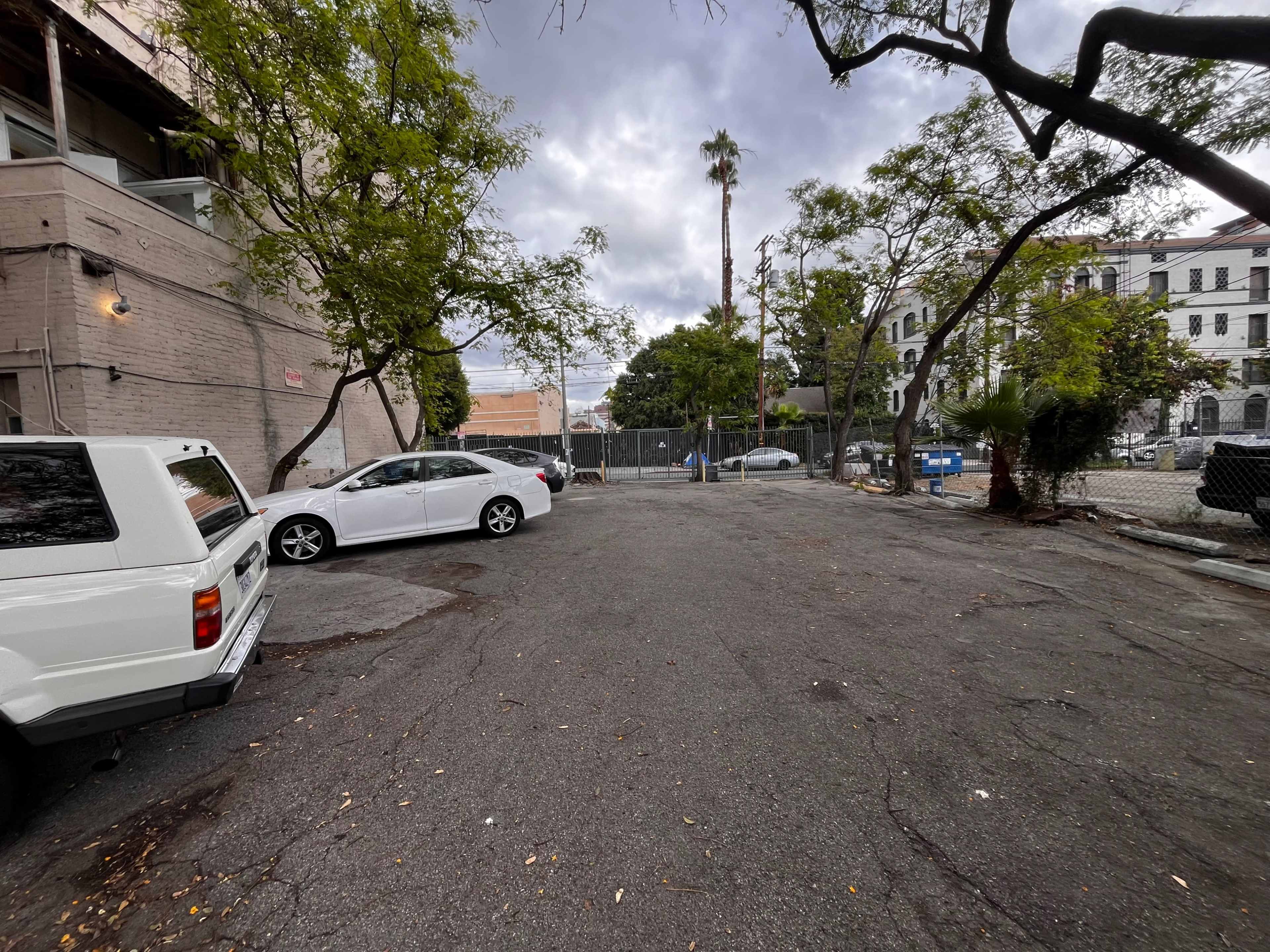 The image depicts a vacant, paved parking lot surrounded by trees and buildings under a cloudy sky.