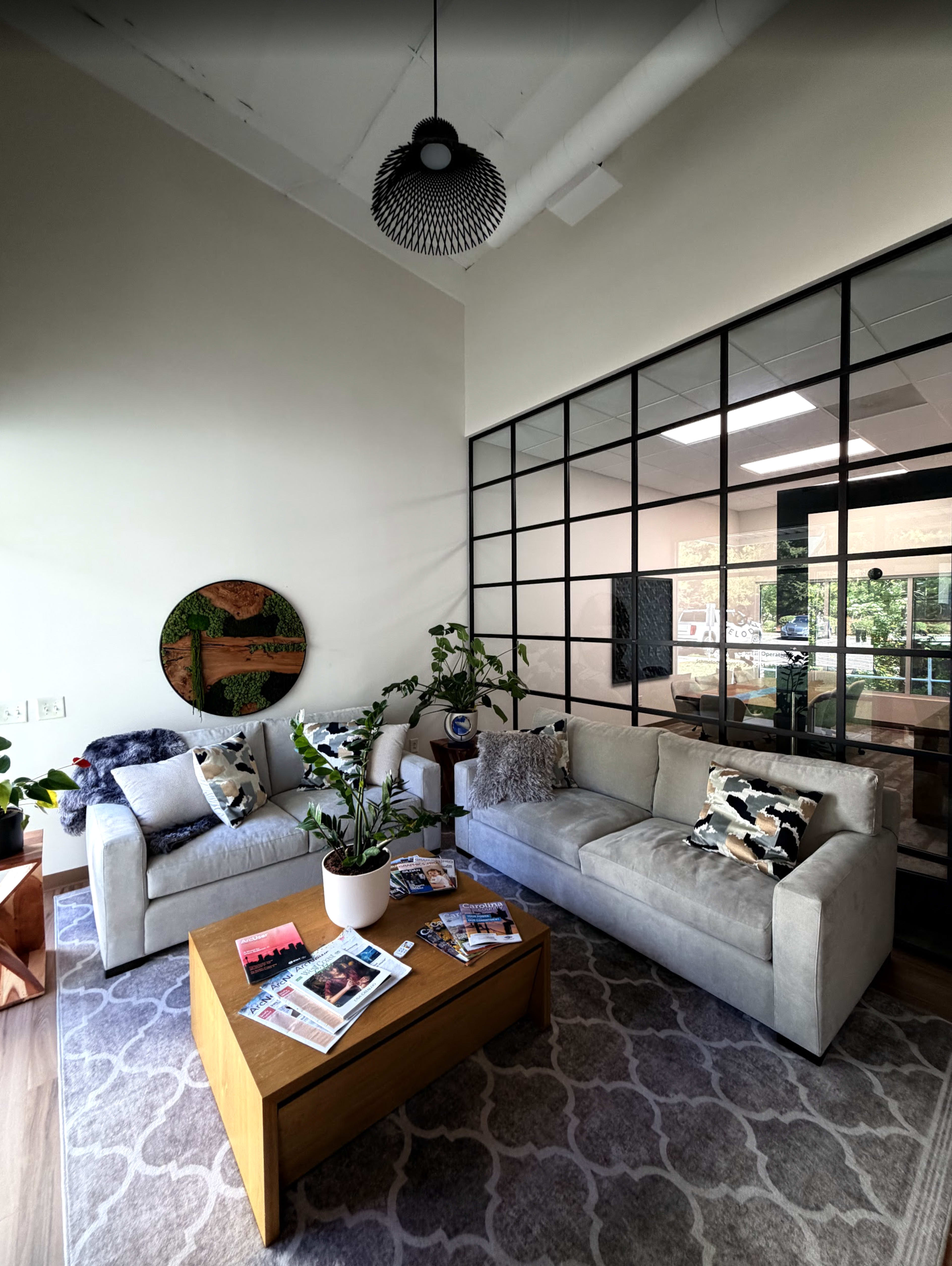 The image shows a modern office lounge with two sofas, a wooden coffee table, various plants, and a decorative wall feature, framed by large glass windows.