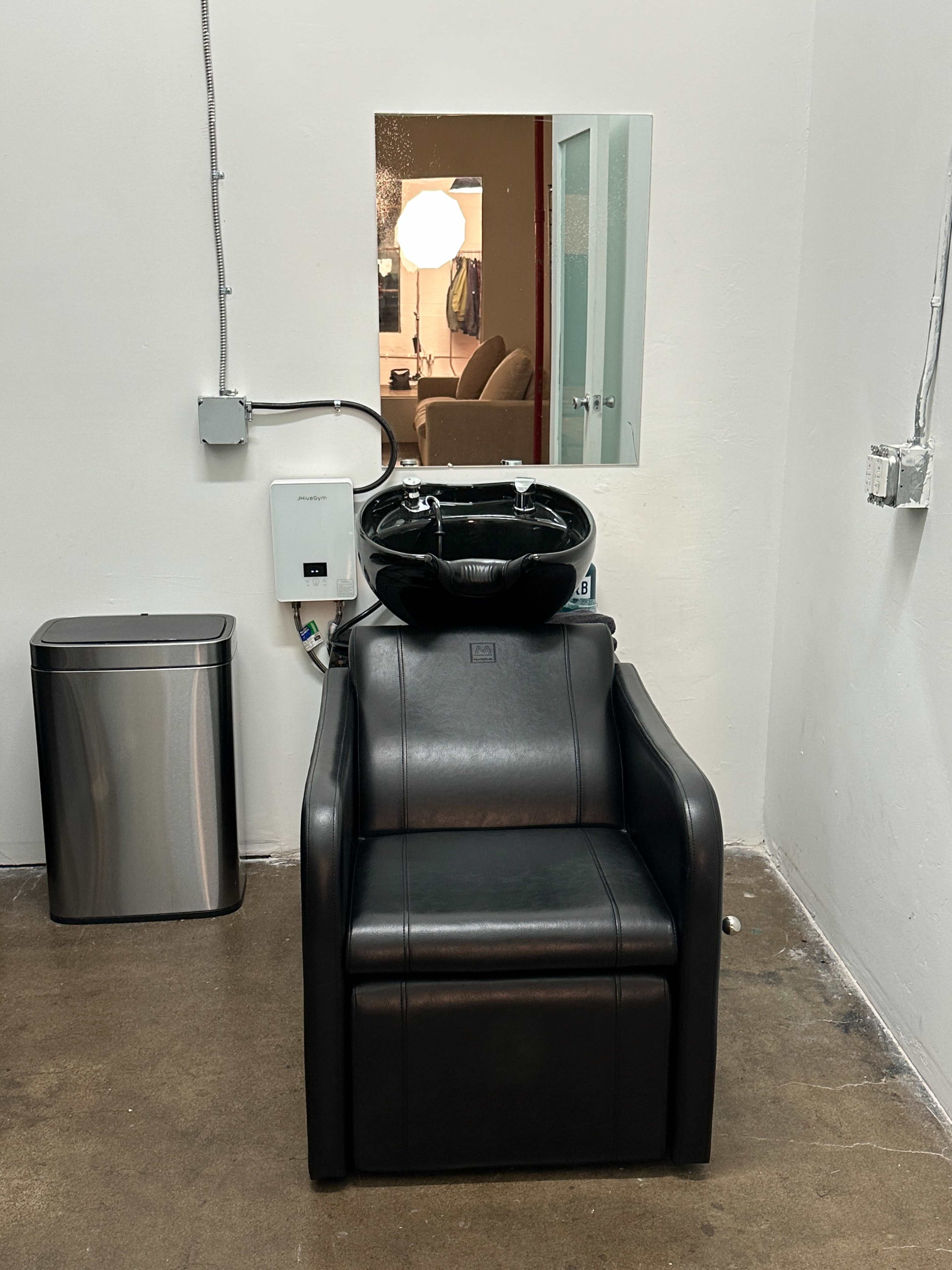 The image shows a black salon chair with a washbasin positioned in front of a mirror in a minimalist room.