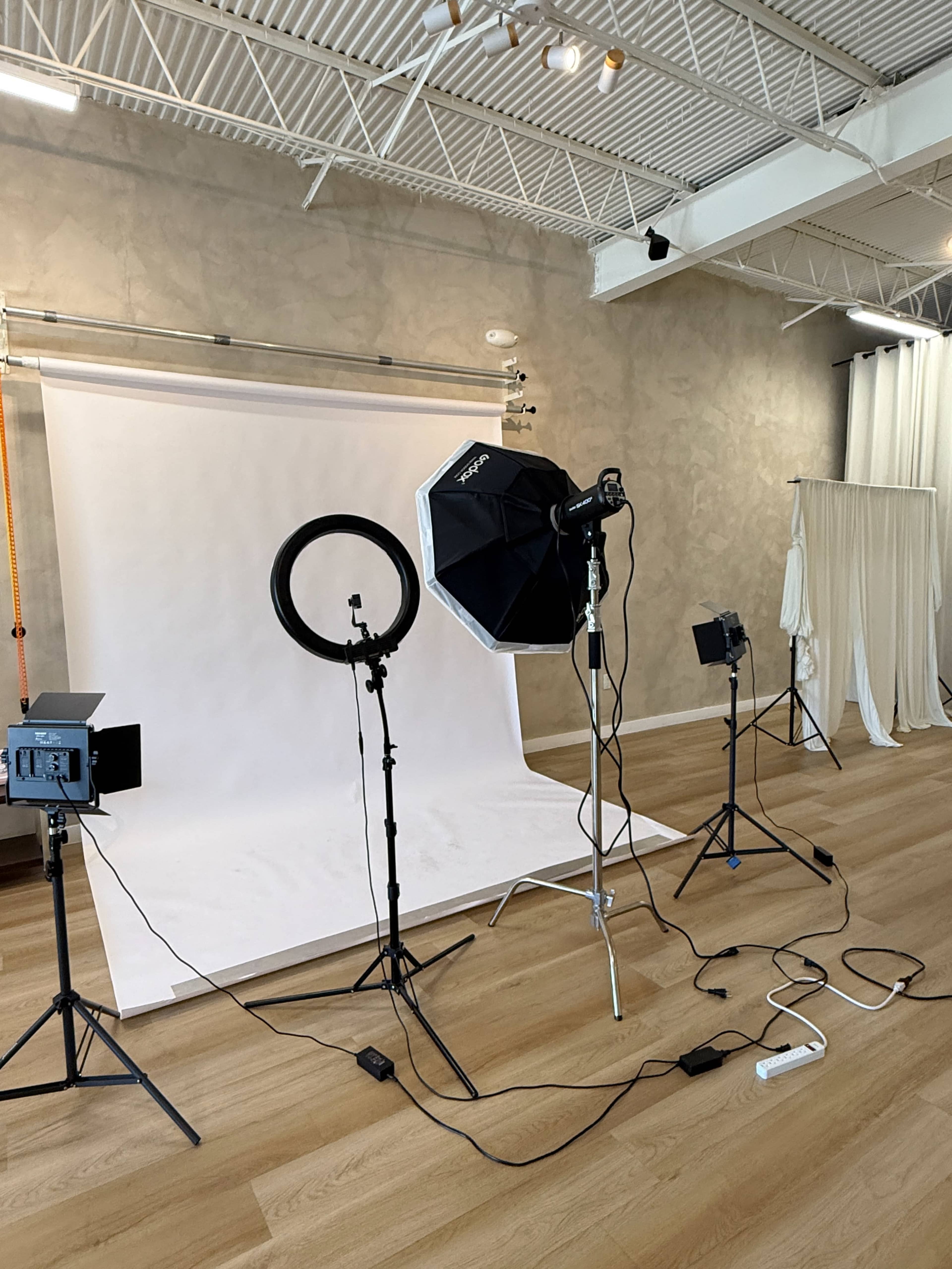 The image shows a photography studio set up with various lighting equipment, including a ring light and softboxes, in front of a white backdrop.