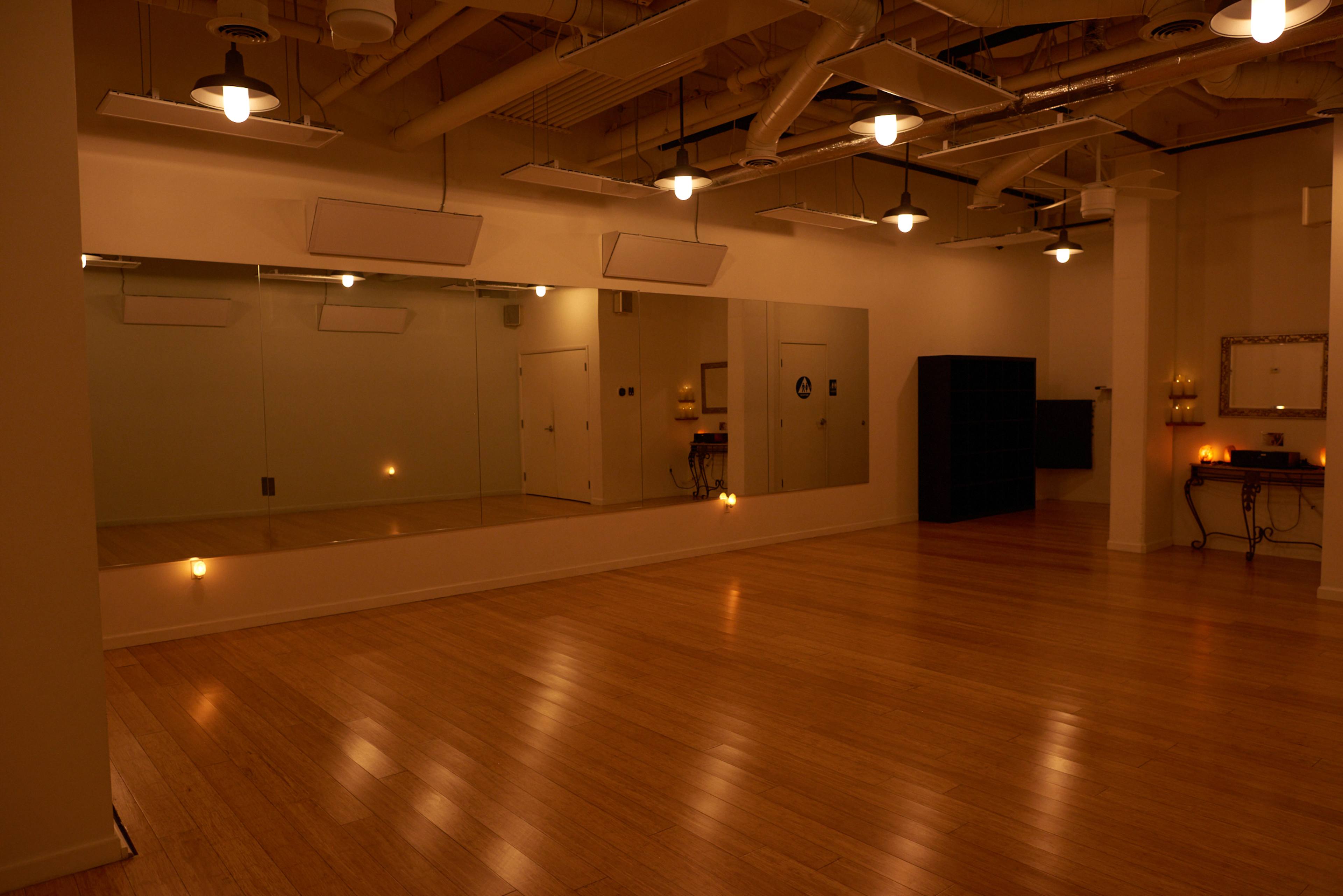 A spacious studio with hardwood flooring and mirrors along one wall, illuminated by overhead lights.
