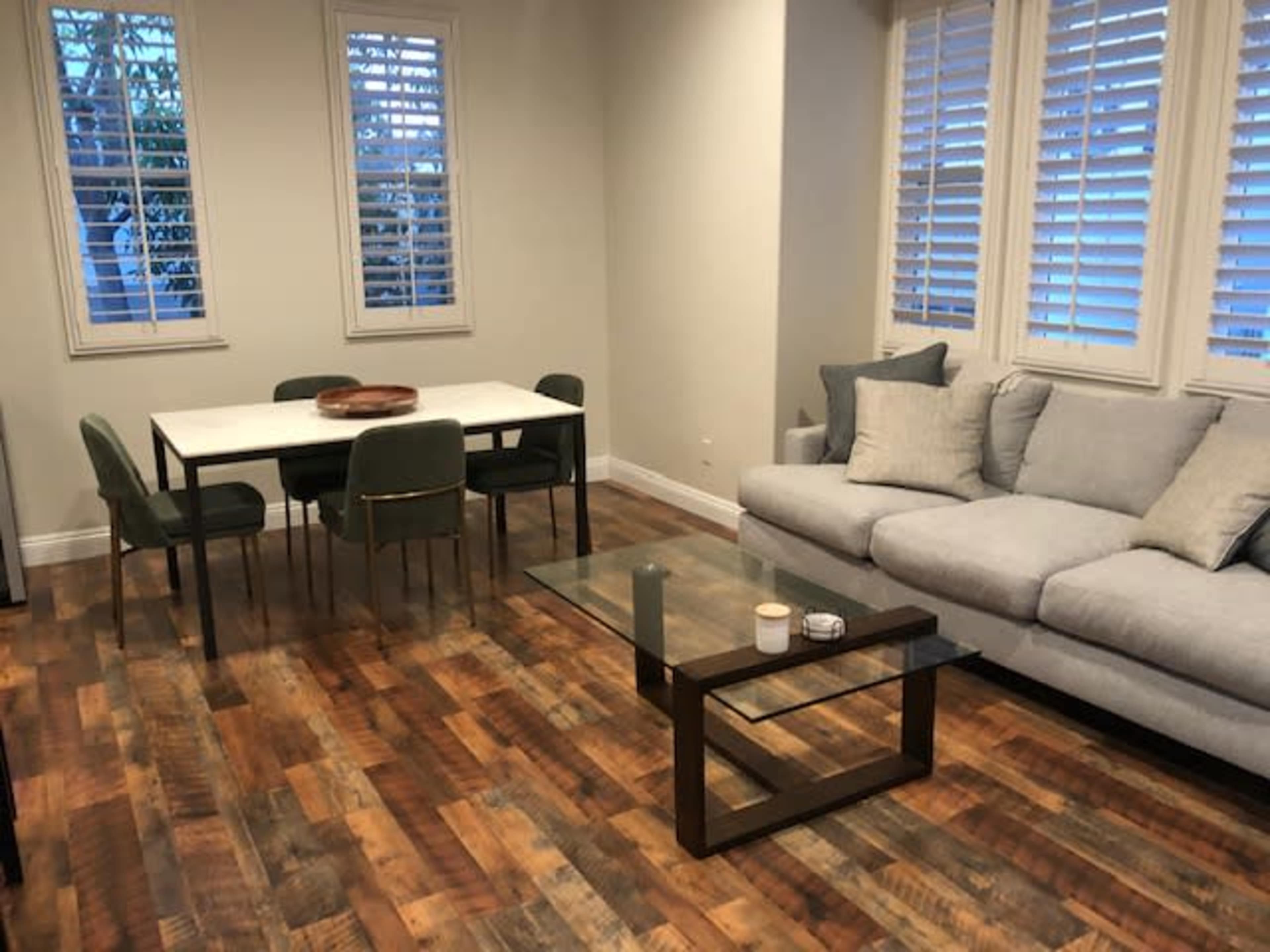 The image shows a room with a gray sofa, a glass coffee table, a white dining table surrounded by green chairs, and large windows with shutters.