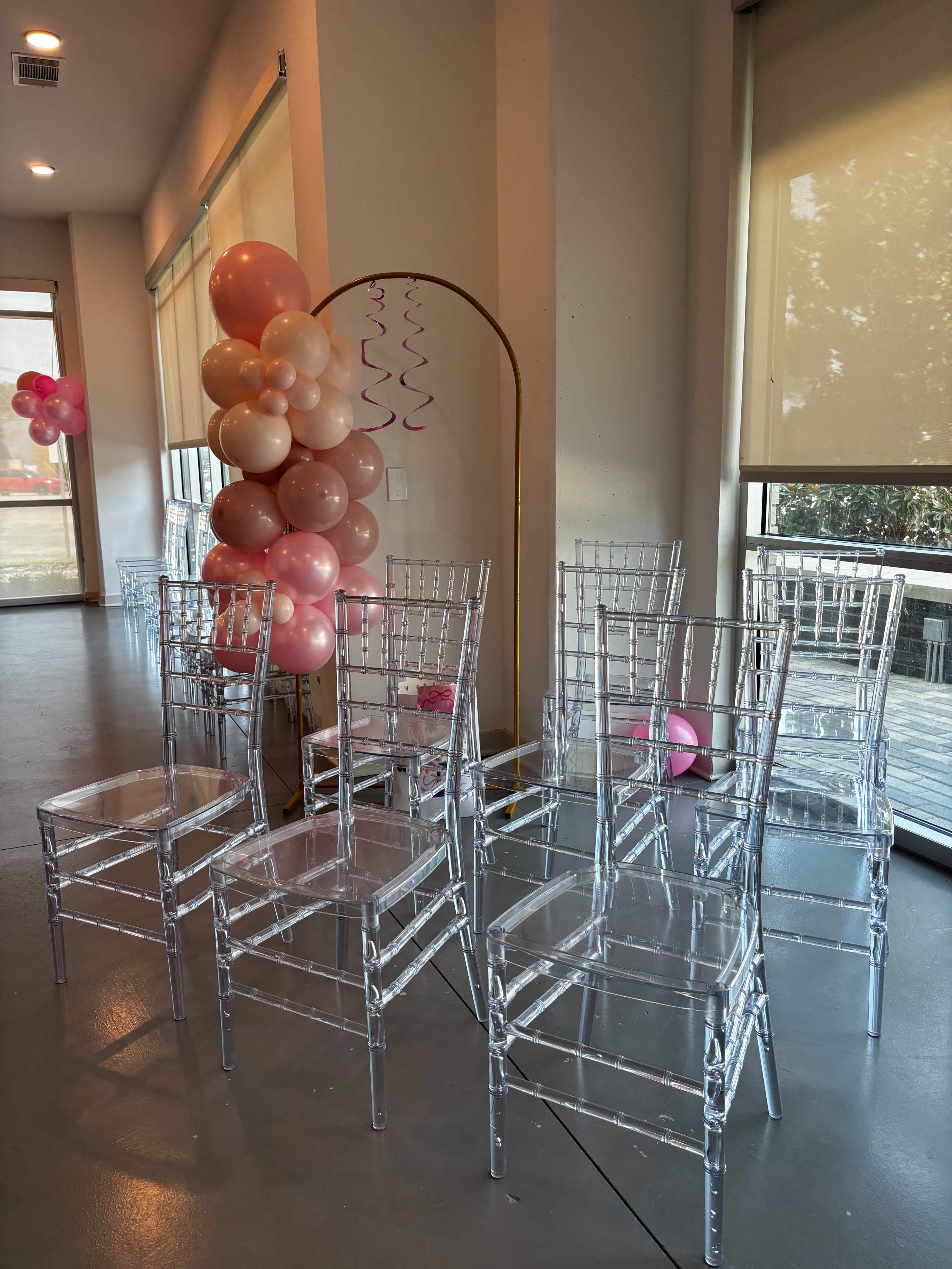 The image shows clear acrylic chairs arranged around a decorated area featuring a cluster of pink and beige balloons and a gold arch in the background.