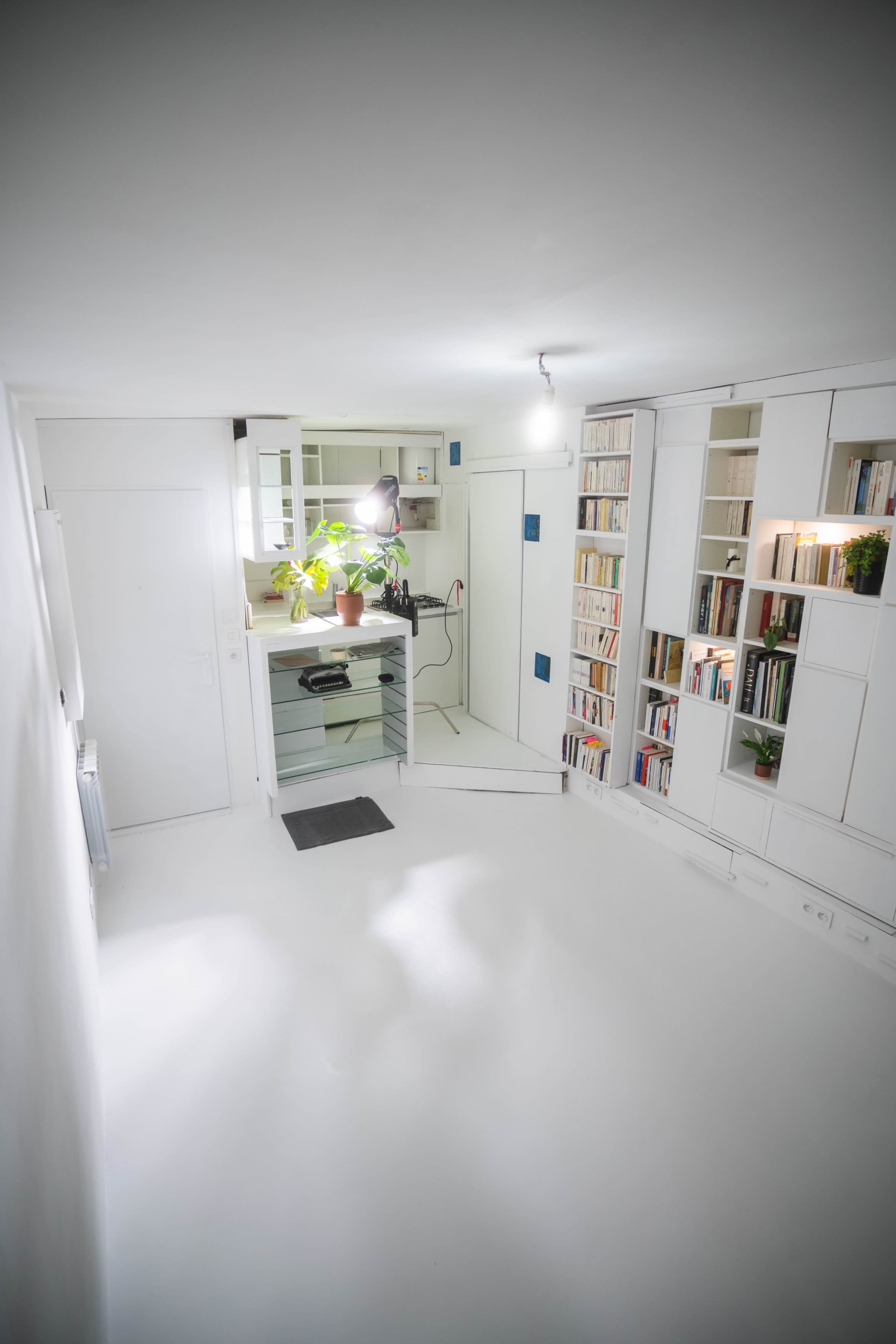 A bright, minimalist room features a small kitchen area with a bookshelf, potted plant, and white walls and flooring.