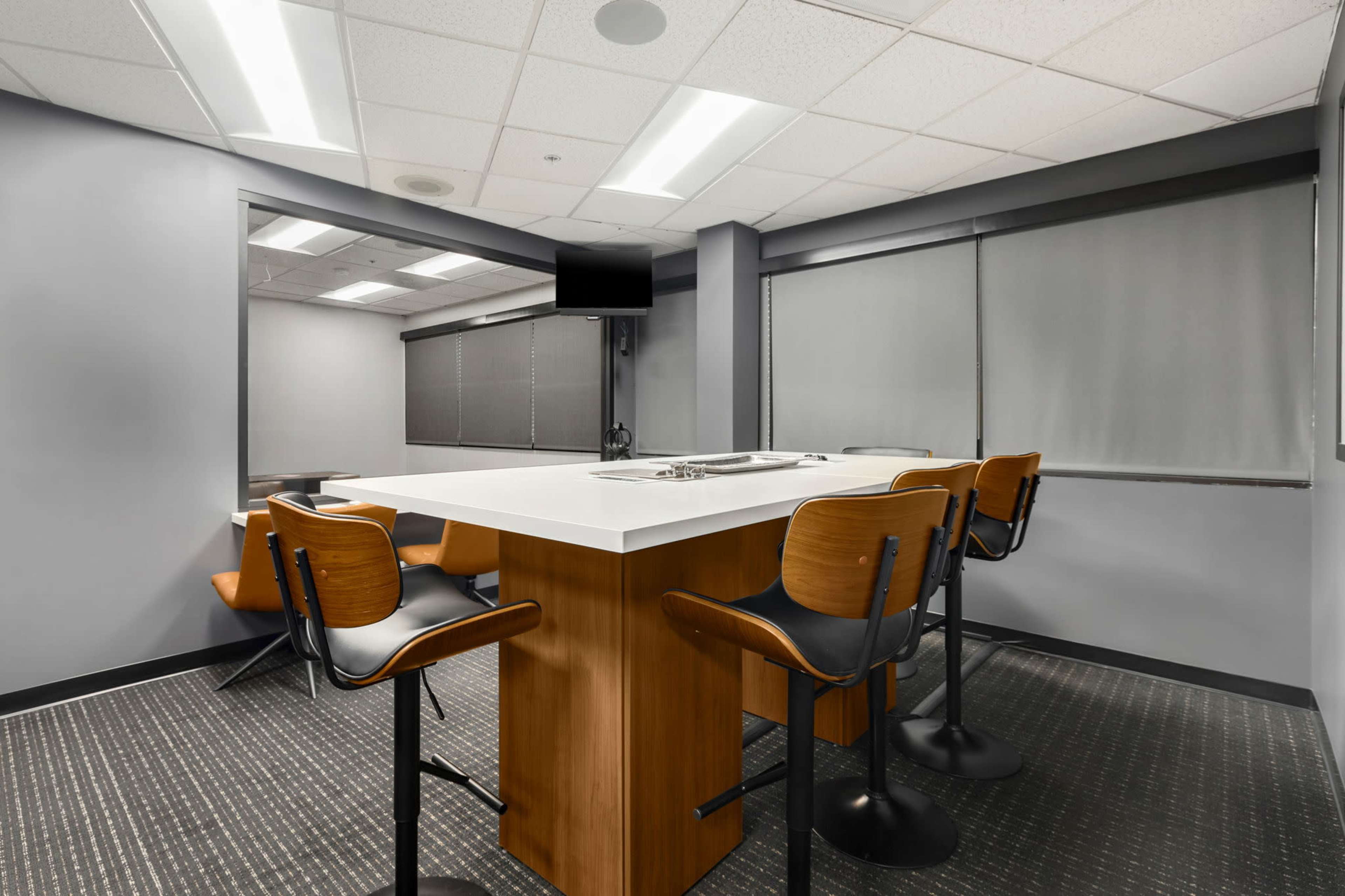 A modern meeting room features a high table with four stools and large windows covered by shades.