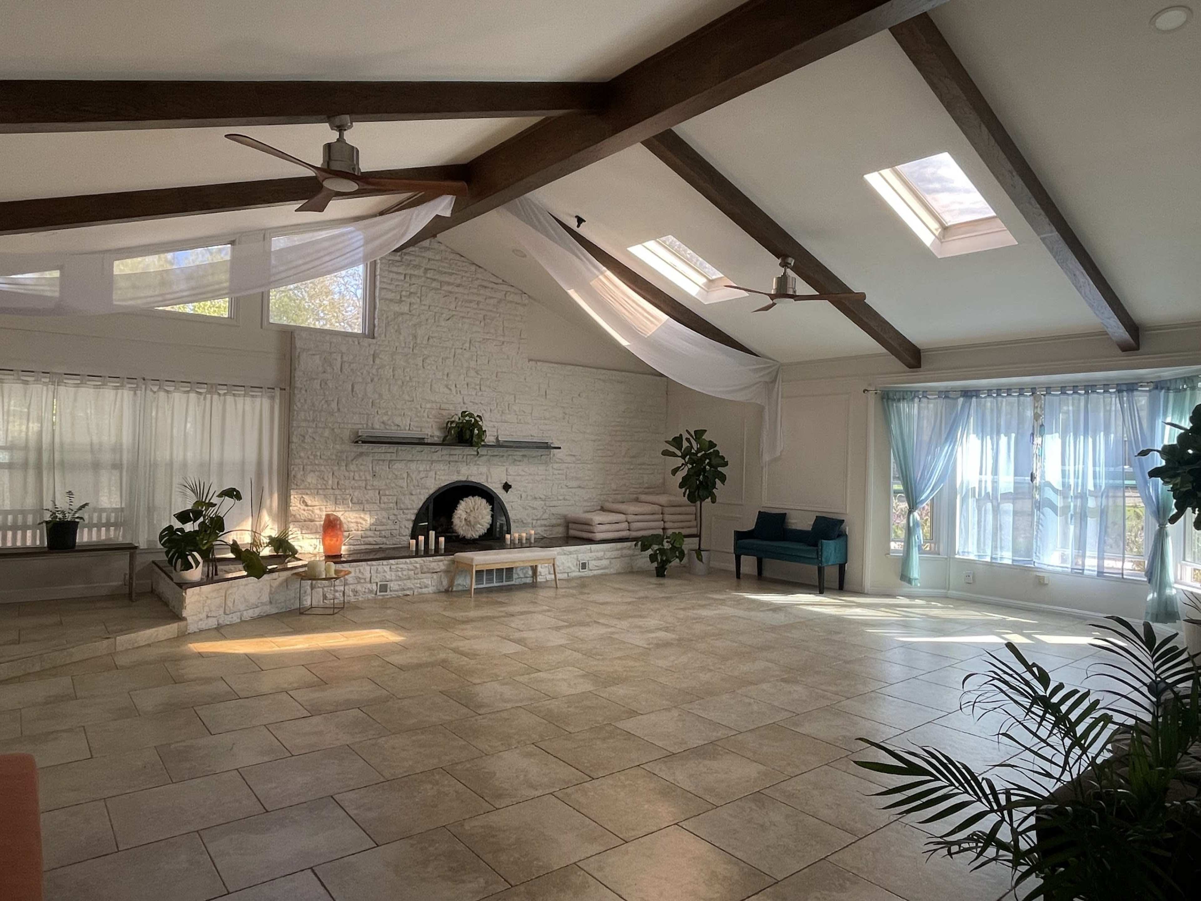The image shows a spacious, well-lit room with high ceilings, large windows, and a stone fireplace, furnished with potted plants and a blue sofa.