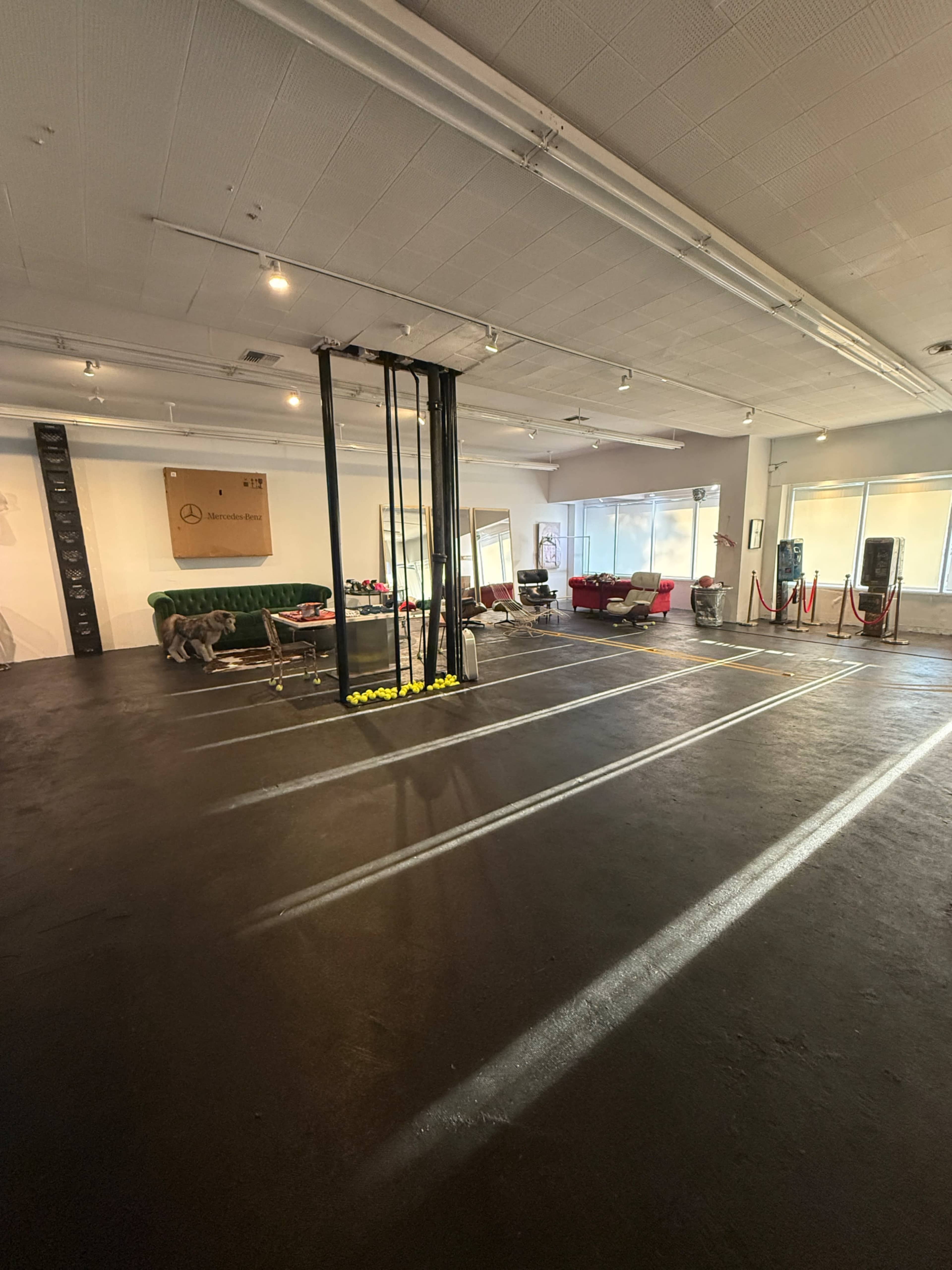 The image shows a spacious indoor area with a green sofa, a red armchair, and a structured frame in the center, surrounded by bright lighting and large windows.