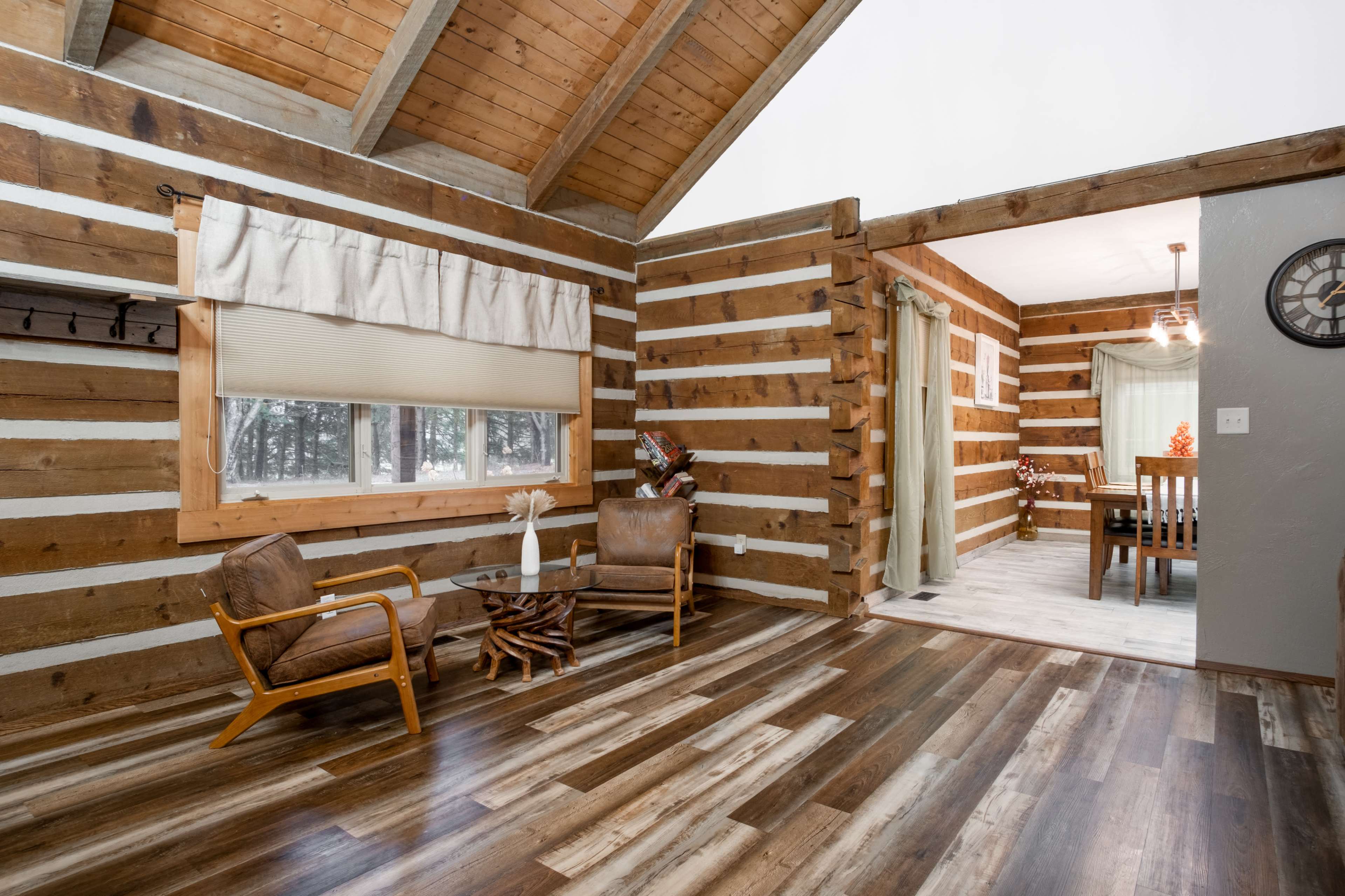 The interior of a log cabin features wooden walls, wooden flooring, and a cozy seating area with a chair and small table, leading to a visible dining area.