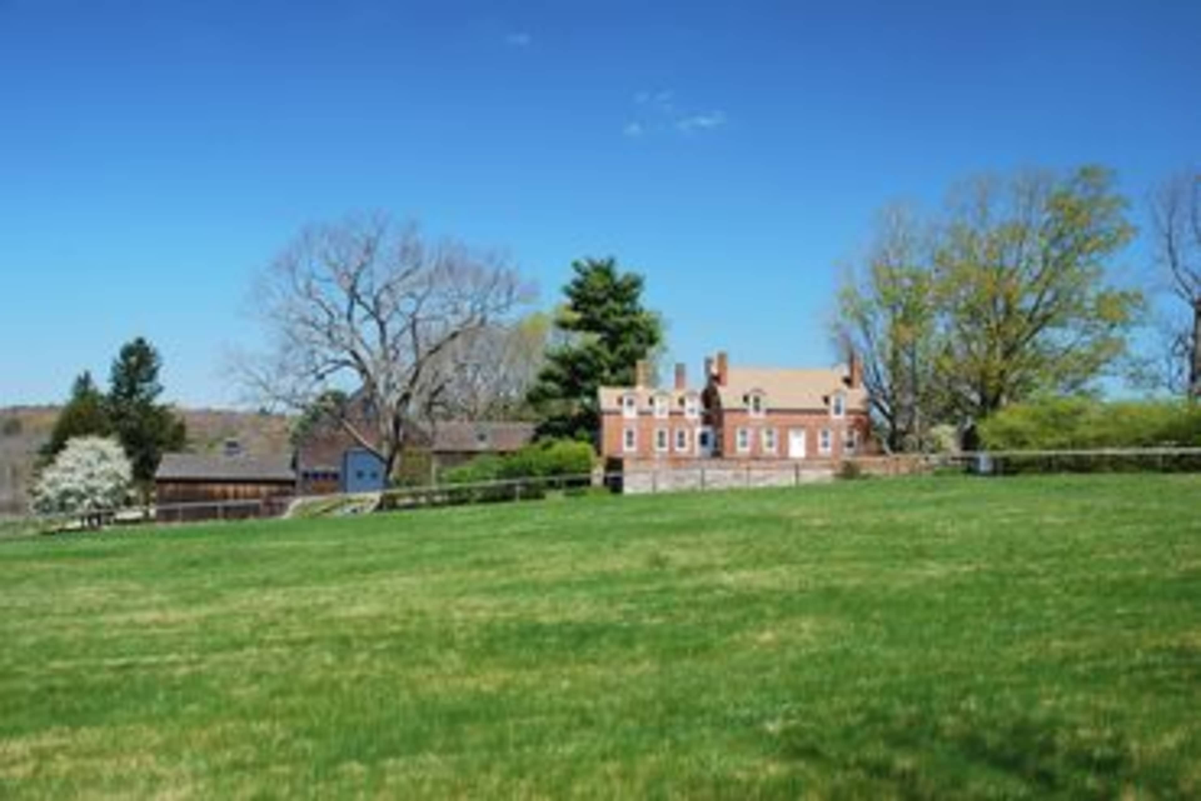 A large brick house stands on a grassy hillside surrounded by trees and outbuildings.