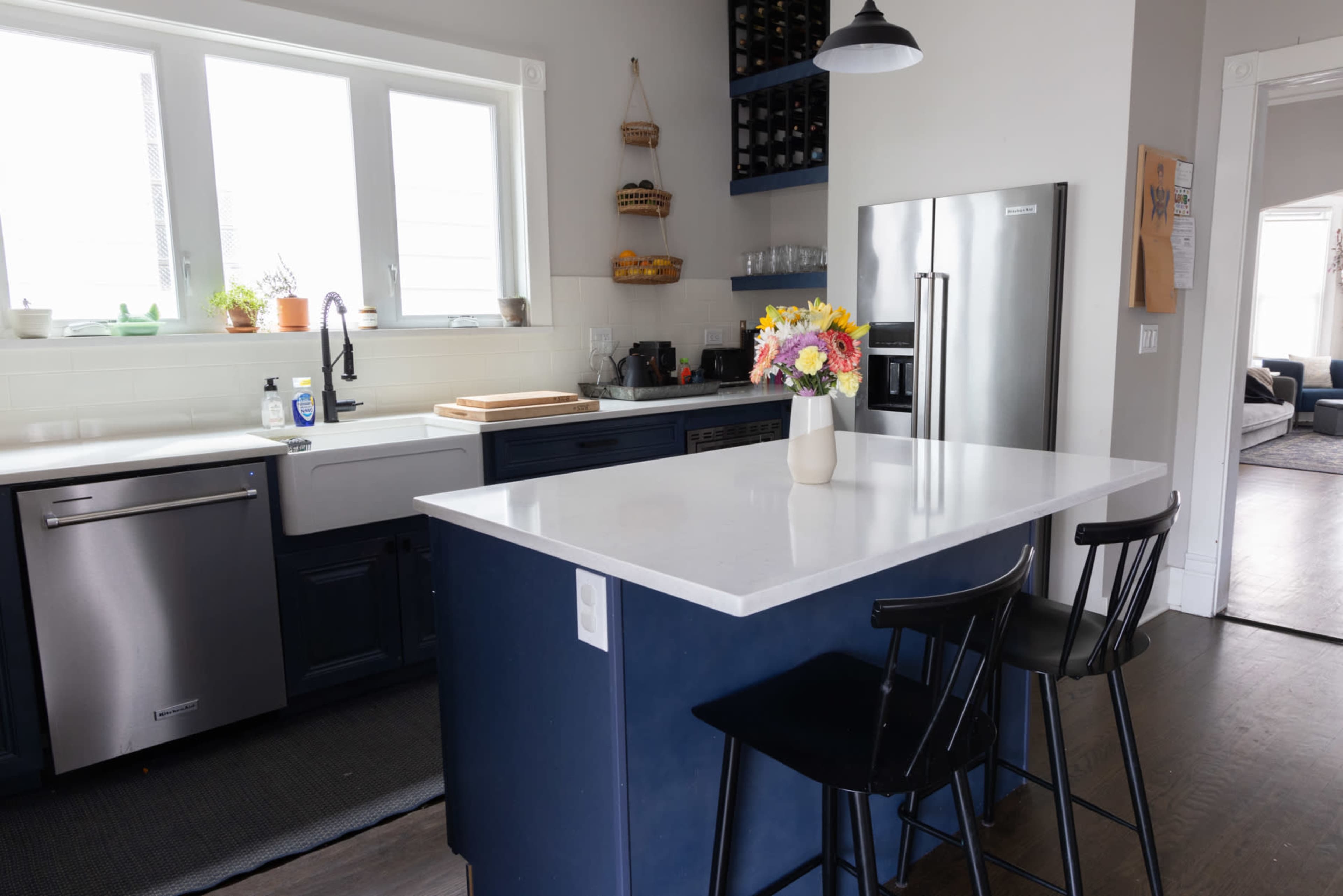 A modern kitchen features a central island with black bar stools, a stainless steel refrigerator, and blue cabinetry, accented by a vase of flowers on the countertop.