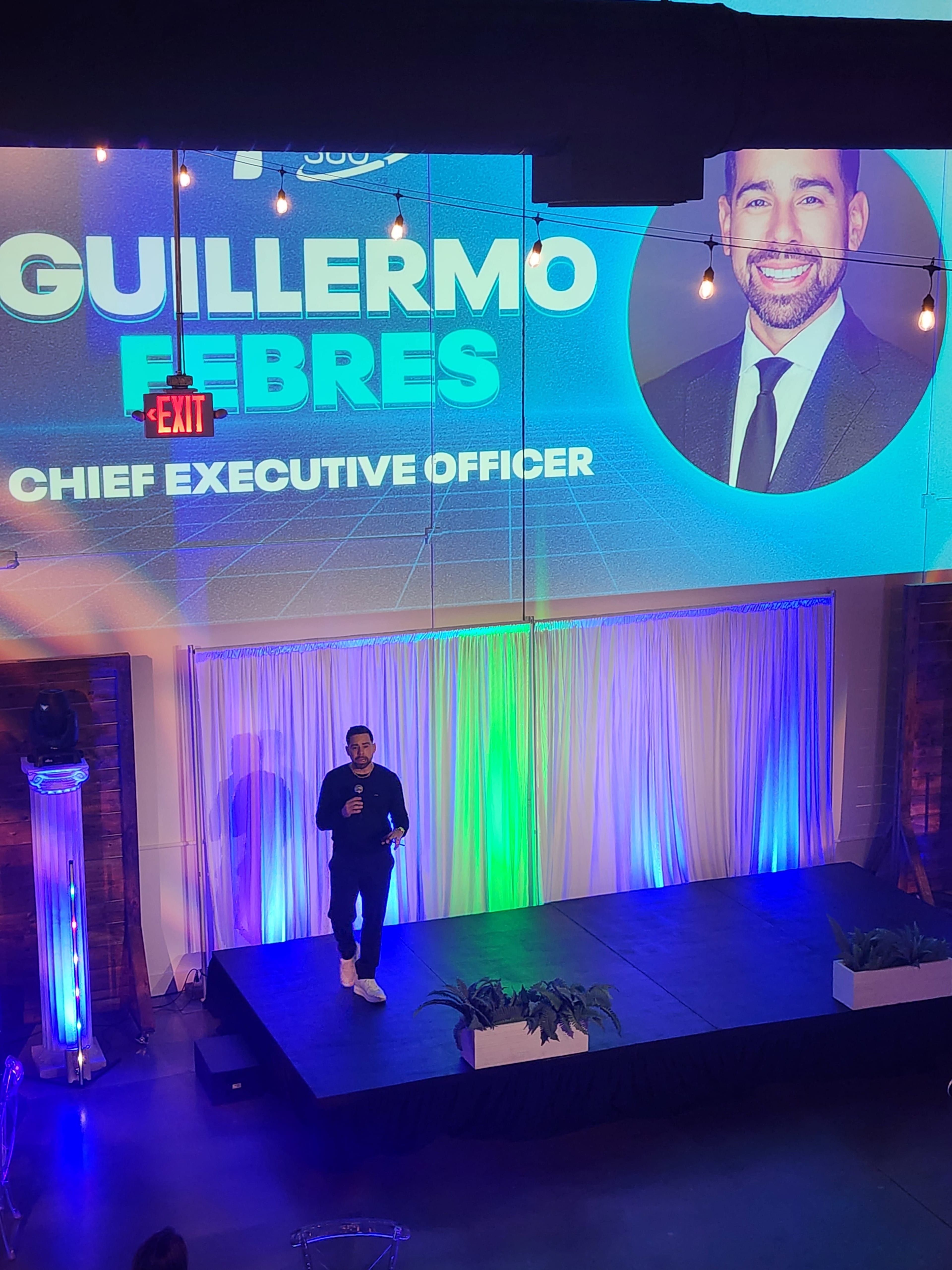 A man stands on a stage speaking, with a large screen behind him displaying his name and title as Chief Executive Officer.