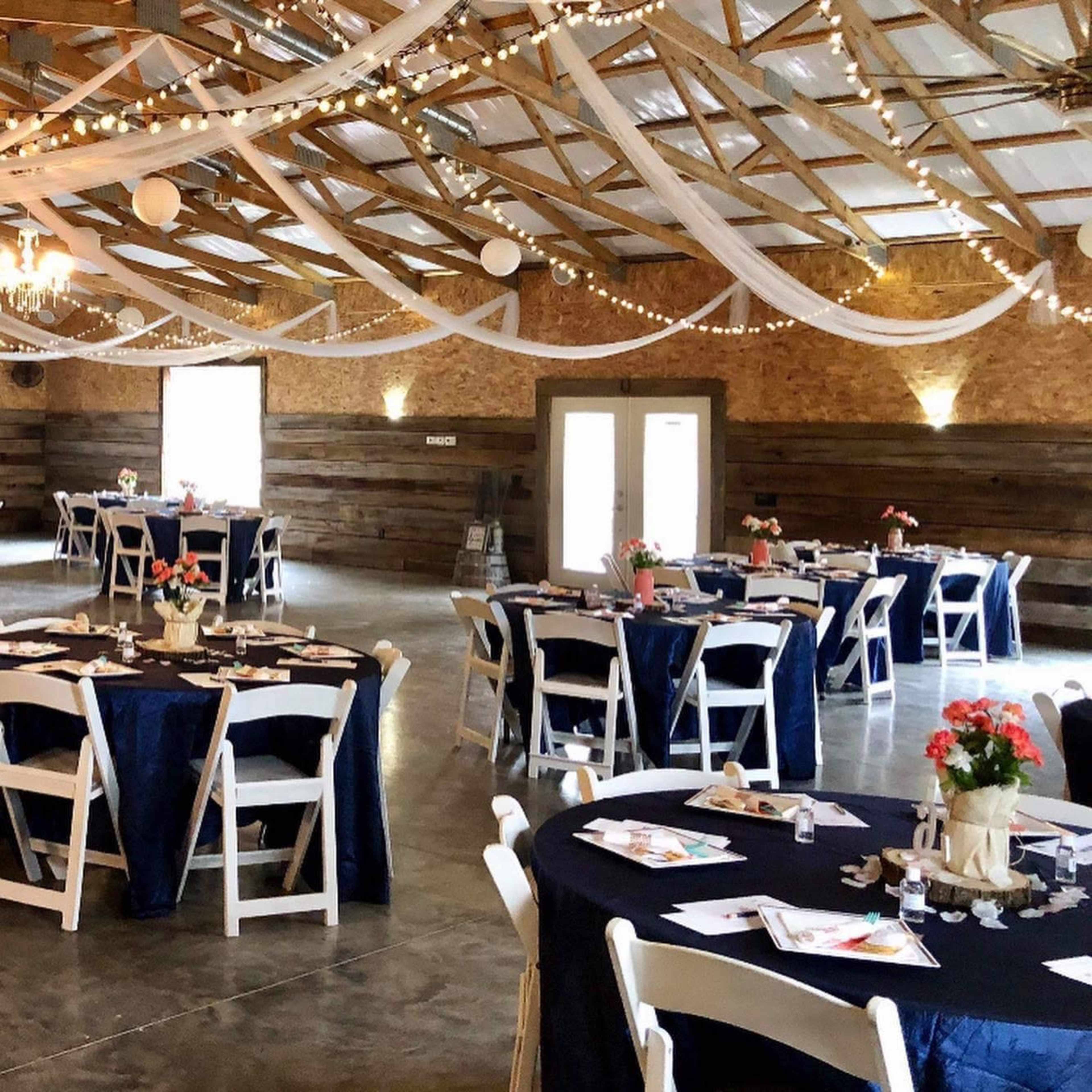 A spacious event venue with several round tables, each set with place settings and floral centerpieces, under a ceiling decorated with draped fabric and string lights.