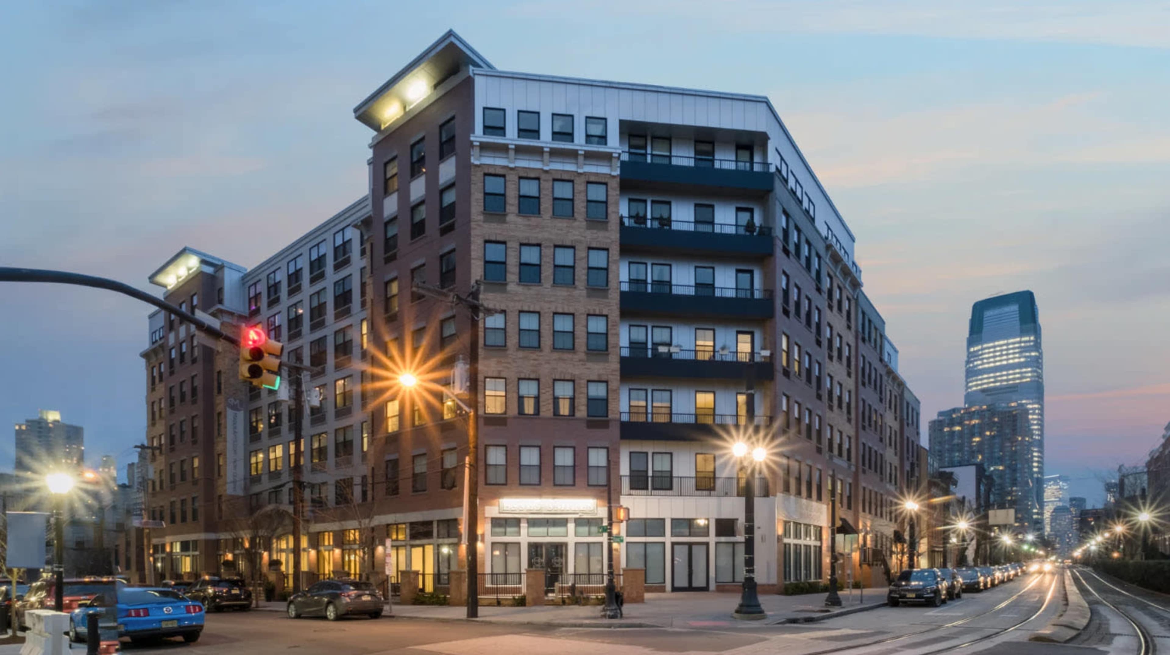 A multi-story residential building with commercial spaces on the ground floor is situated at a street intersection, illuminated by streetlights and traffic signals, with a city skyline visible in the background at dusk.