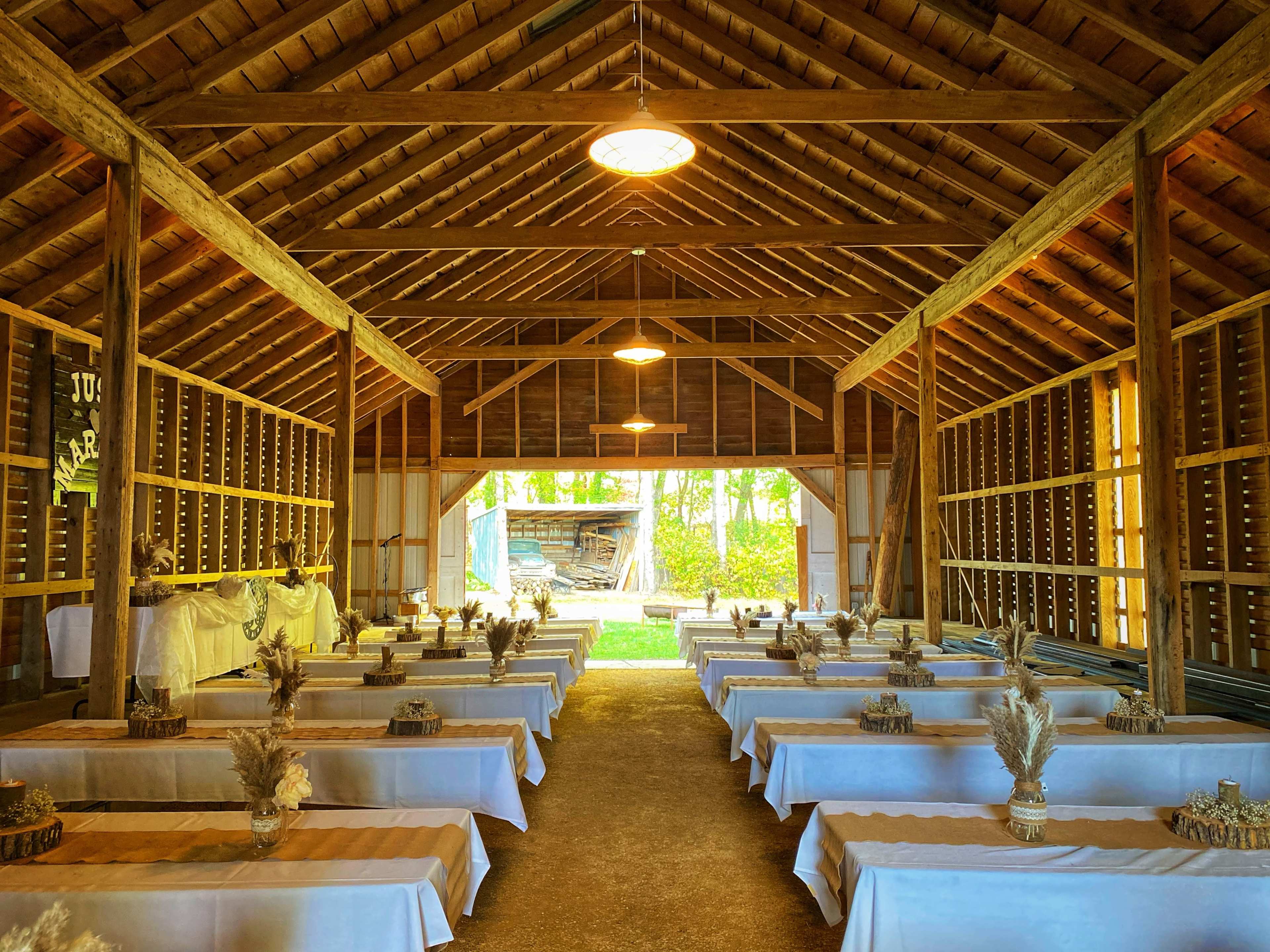 The interior of a rustic barn features rows of tables dressed in white cloth, arranged for an event, with wooden beams and natural light streaming in from an open doorway.