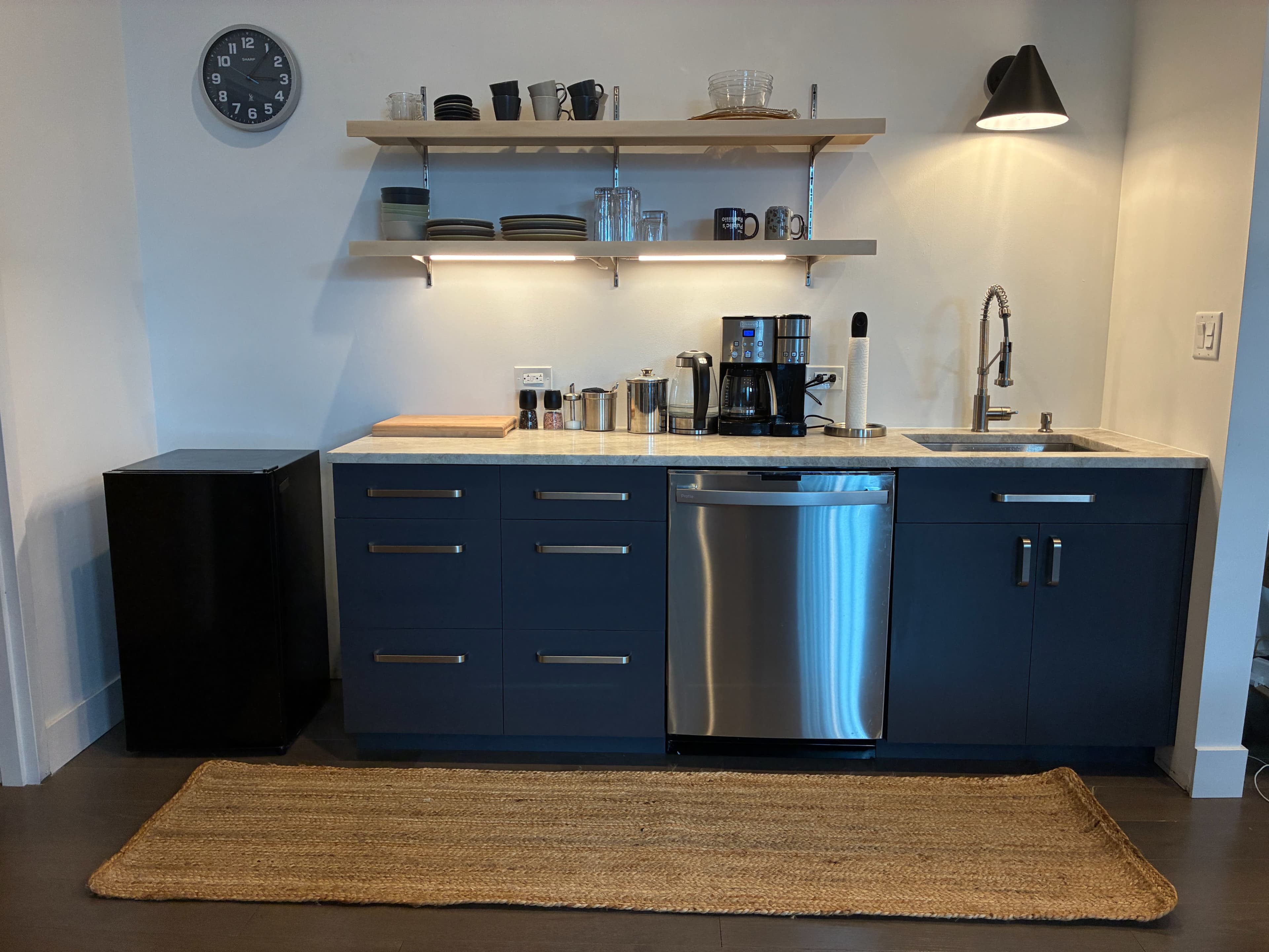 The image shows a modern kitchen setup with a dark blue cabinet, a stainless steel dishwasher, a sink, a countertop with coffee equipment, and shelving displaying glassware.