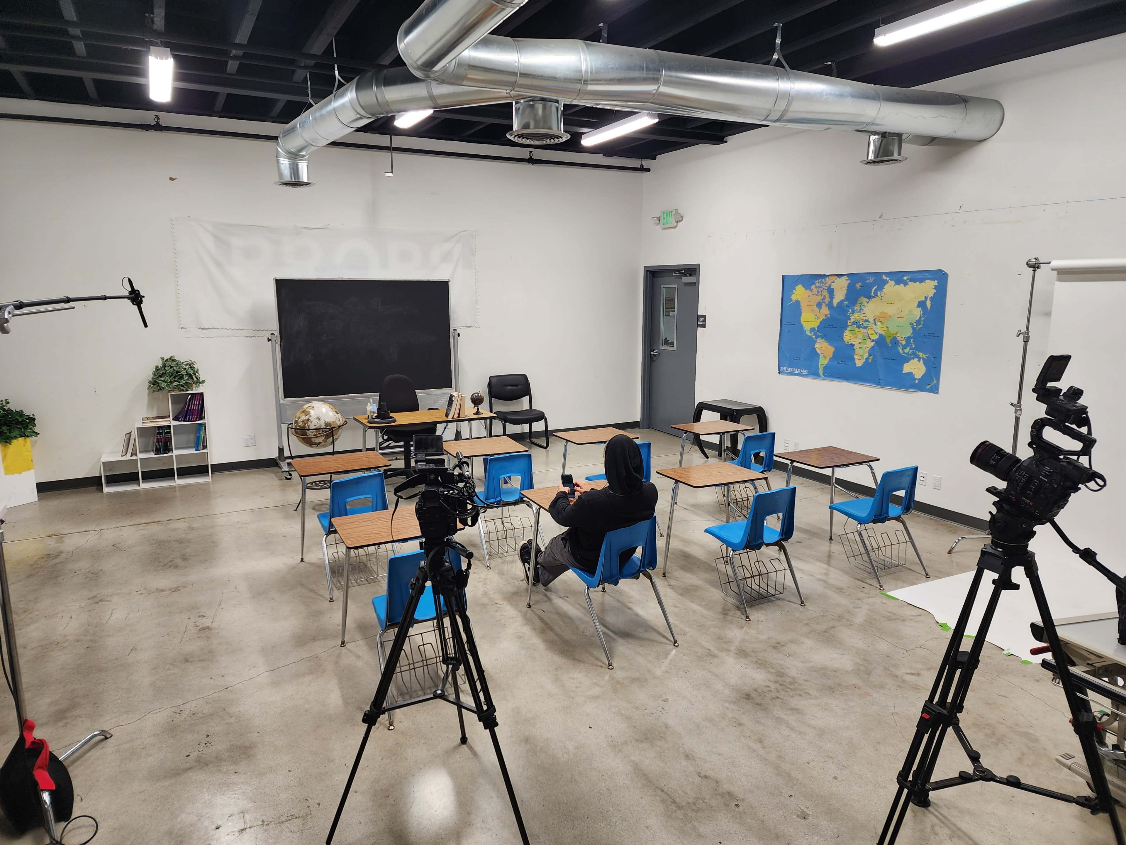 An empty classroom setup with several desks and chairs arranged around a central space, flanked by cameras on tripods.
