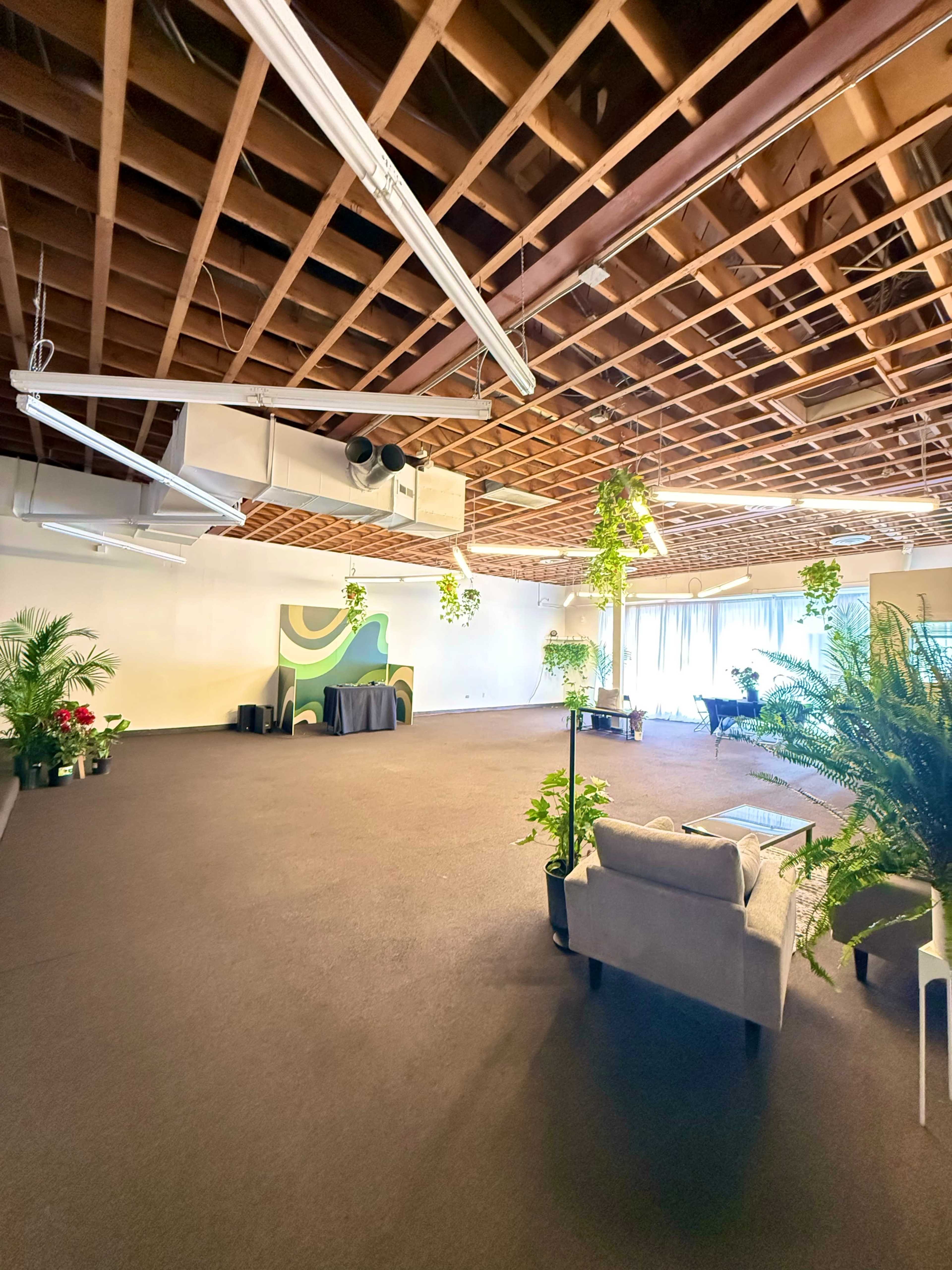 The image shows a spacious room with a designed stage area in front, surrounded by potted plants and a seating arrangement.