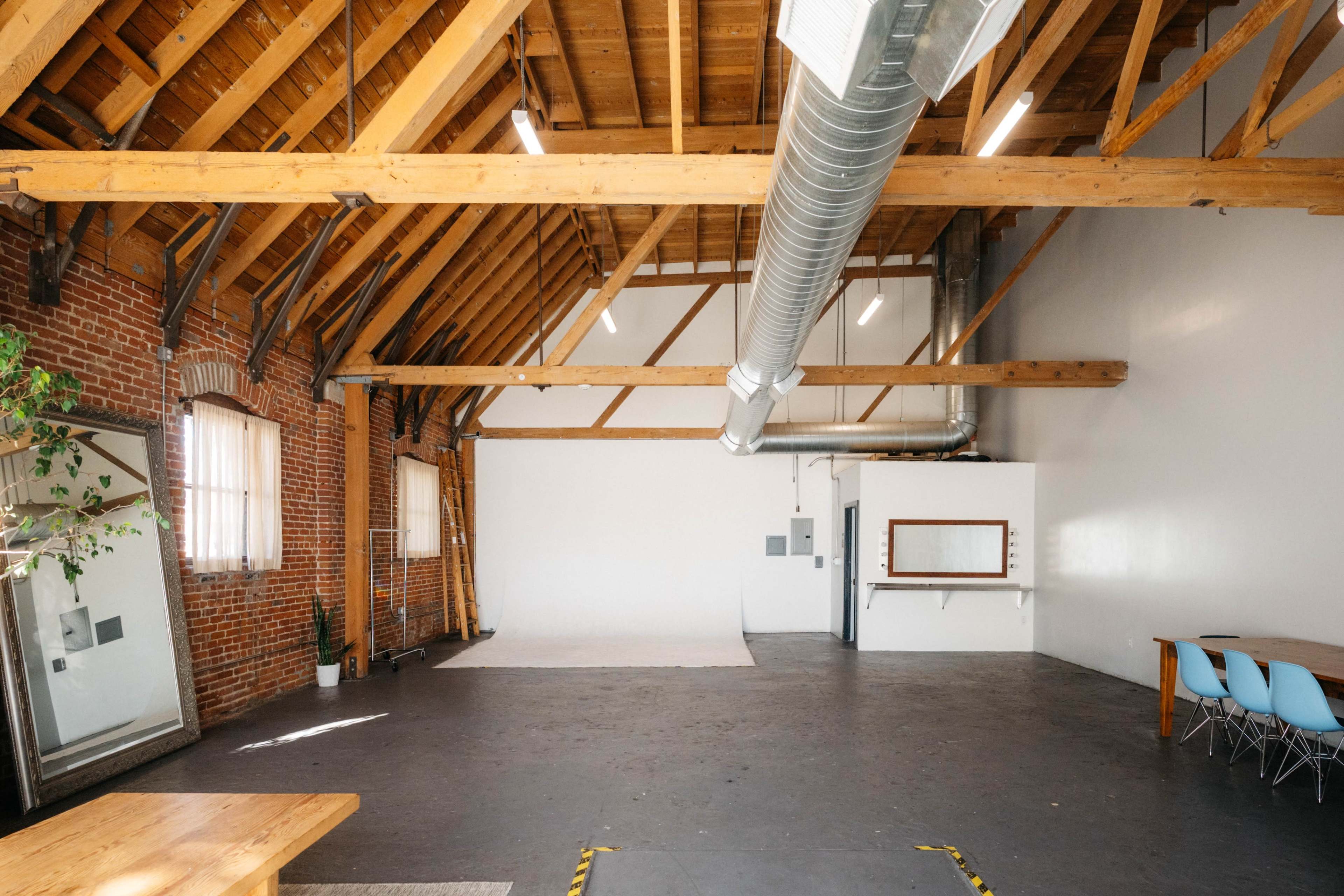 The image shows a spacious loft-style interior with exposed wooden beams, a brick wall, and an empty area featuring a gray floor and large windows.
