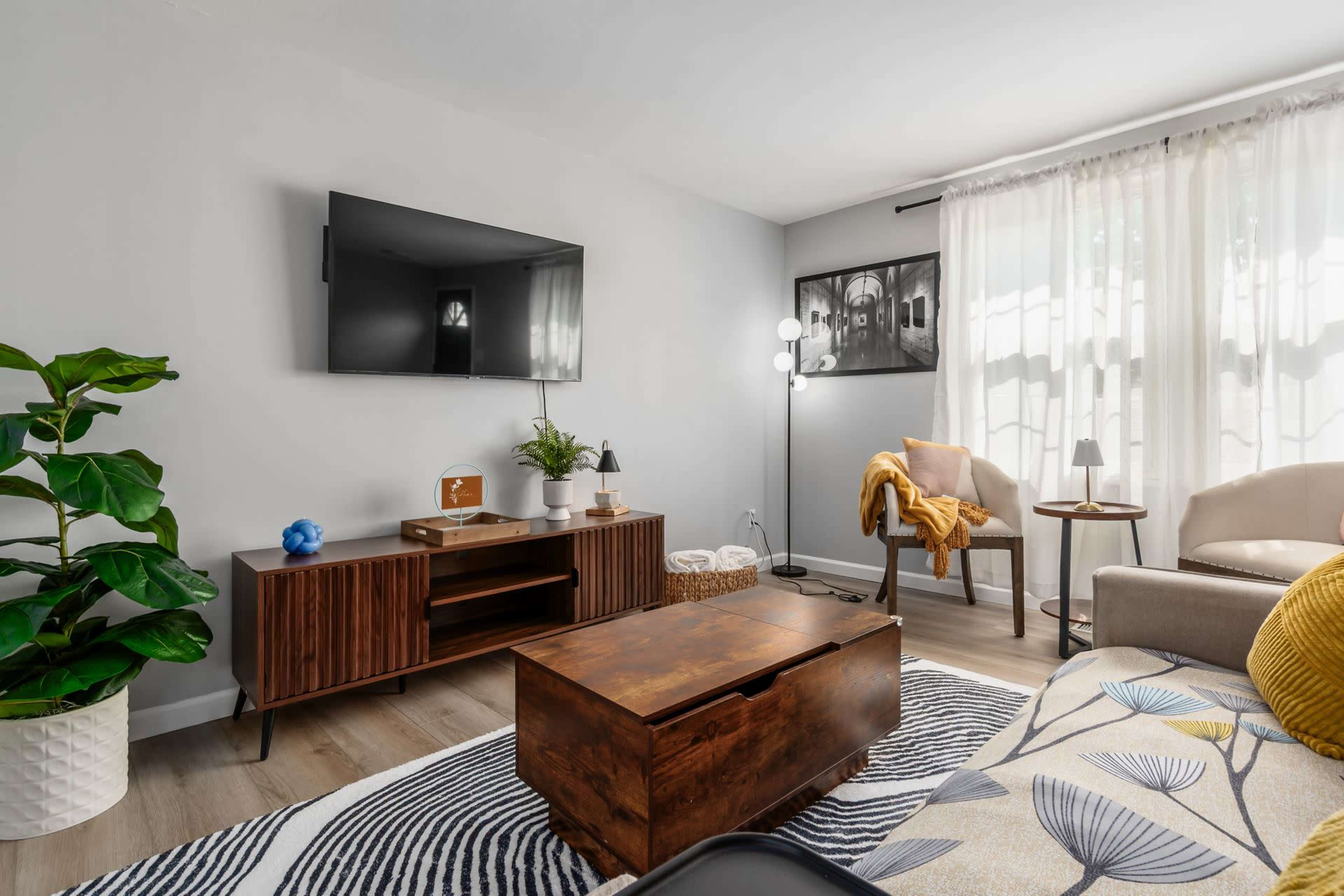 A modern living room features a wall-mounted TV, a wooden coffee table, and a mix of plants, textiles, and furniture arranged around a natural color scheme.
